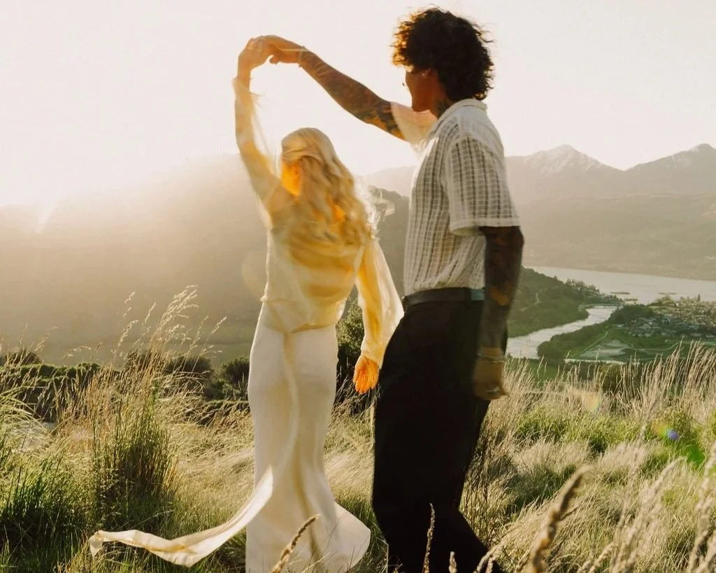 A couple dancing in a field with mountains and a river in the background during sunset.