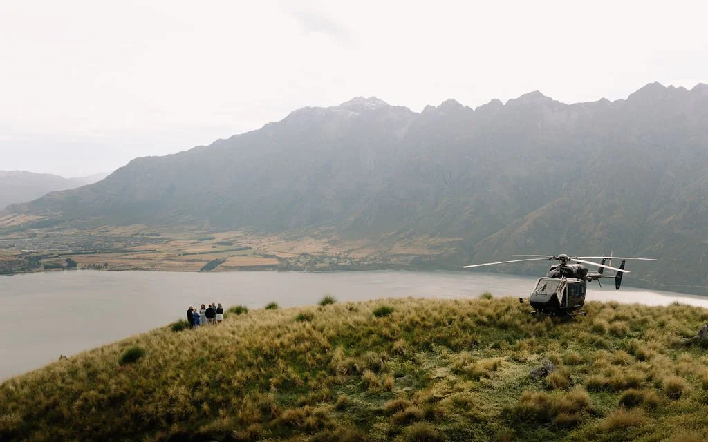 A helicopter parked on a grassy hill overlooking a lake and mountain range, with a group of people near the edge of the hill.