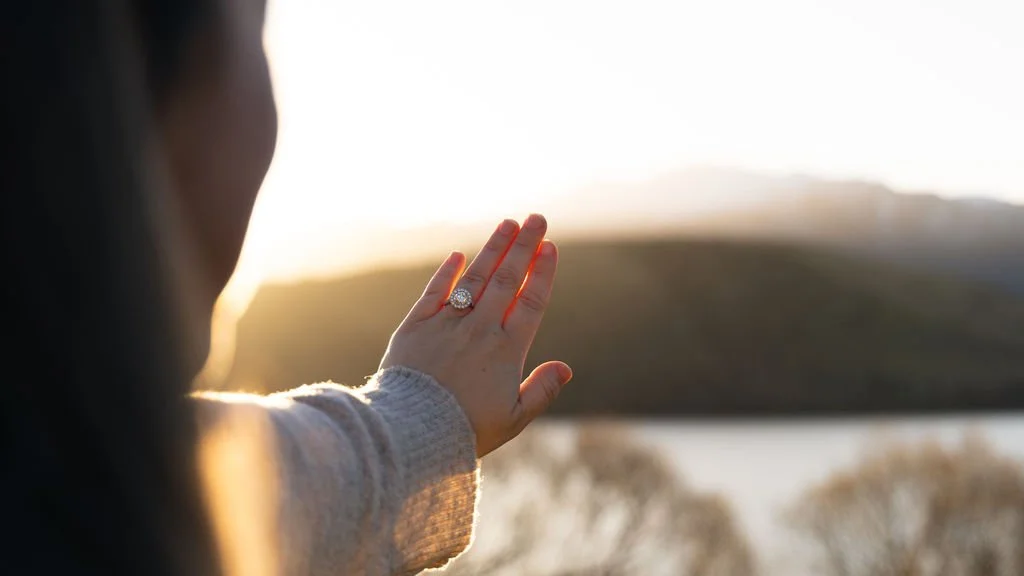 Woman's hand with an engagement ring, facing a sunset over a lake and hills.