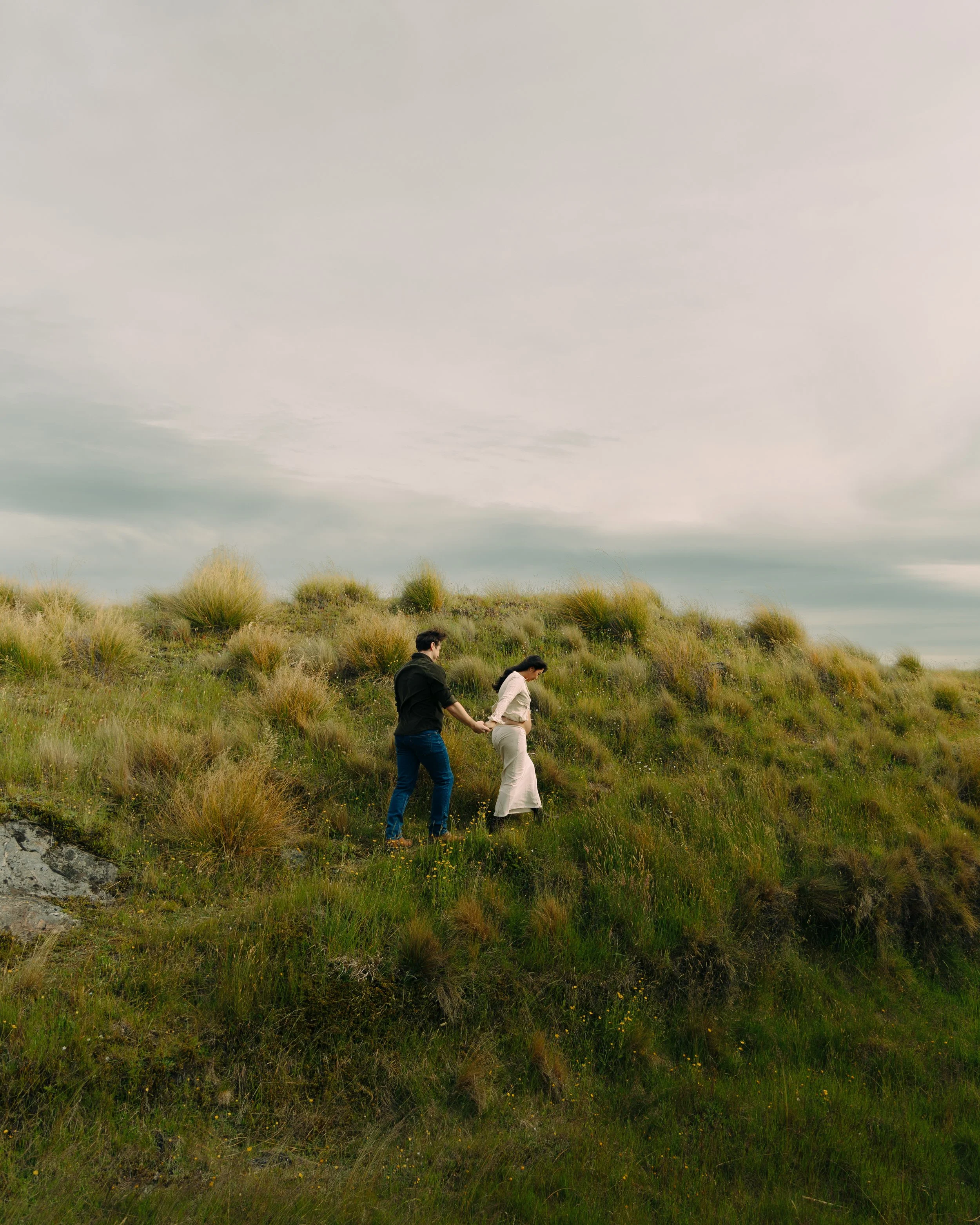 A couple holding hands and walking through grassy dunes under a cloudy sky.