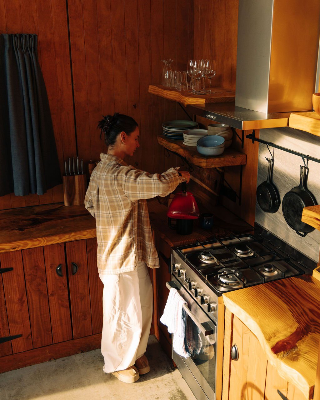 A woman cooking in a rustic kitchen with wood cabinets, open shelves, and hanging cast iron pans.