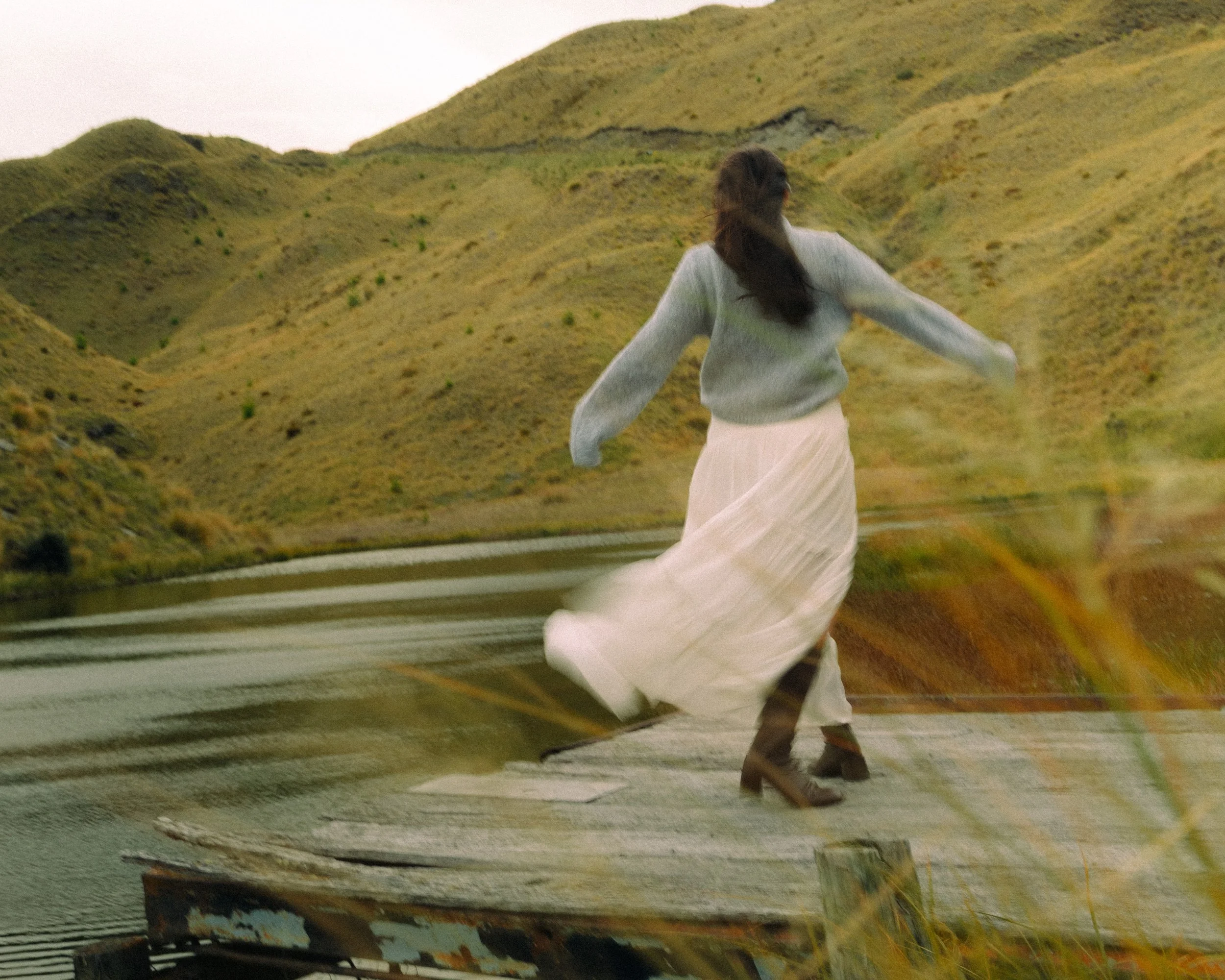 A woman in a gray sweater and long white skirt is running along a rural road bordered by grassy hills.