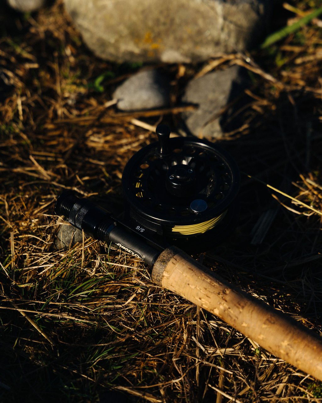 A black fishing reel with a beige handle lying on ground with dry grass, twigs, and rocks.