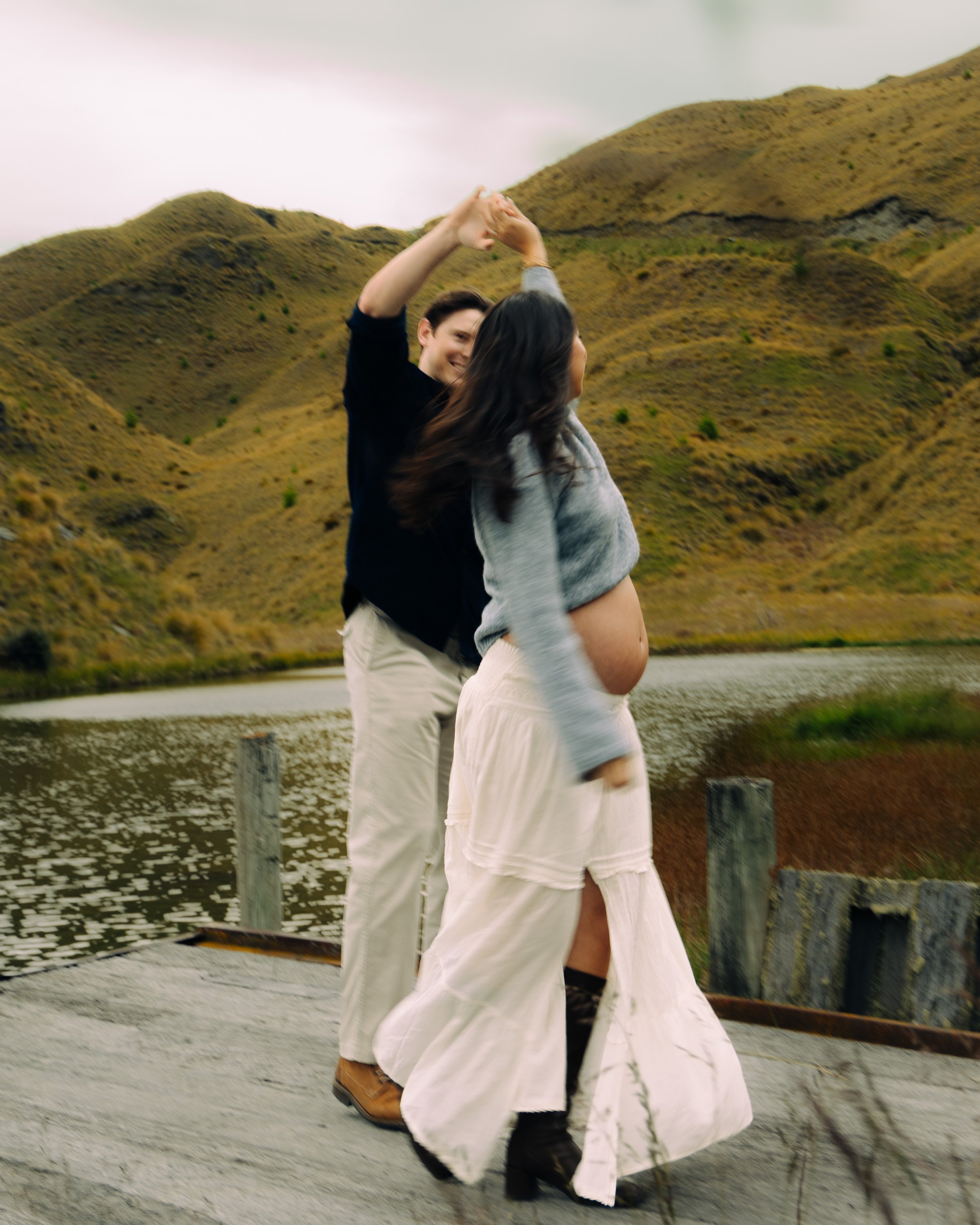 A couple dancing outdoors near a lake with mountains in the background. The woman has a baby bump and is wearing a long cream-colored skirt and black boots. The man is dressed in a black shirt and beige pants.