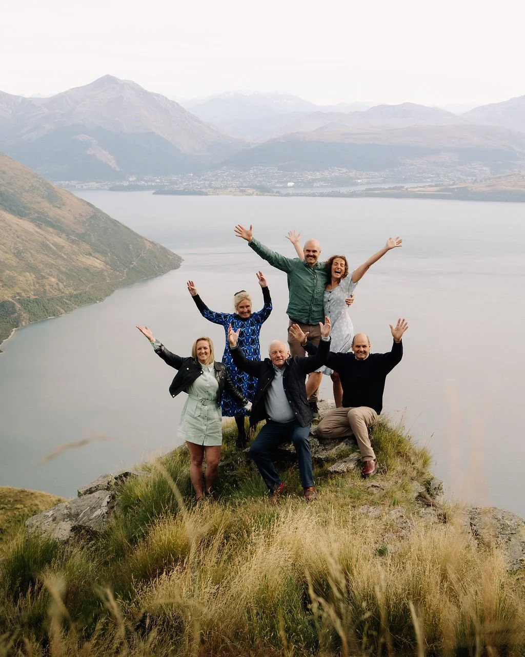 Group of six people celebrating on a hilltop overlooking a lake and mountains.