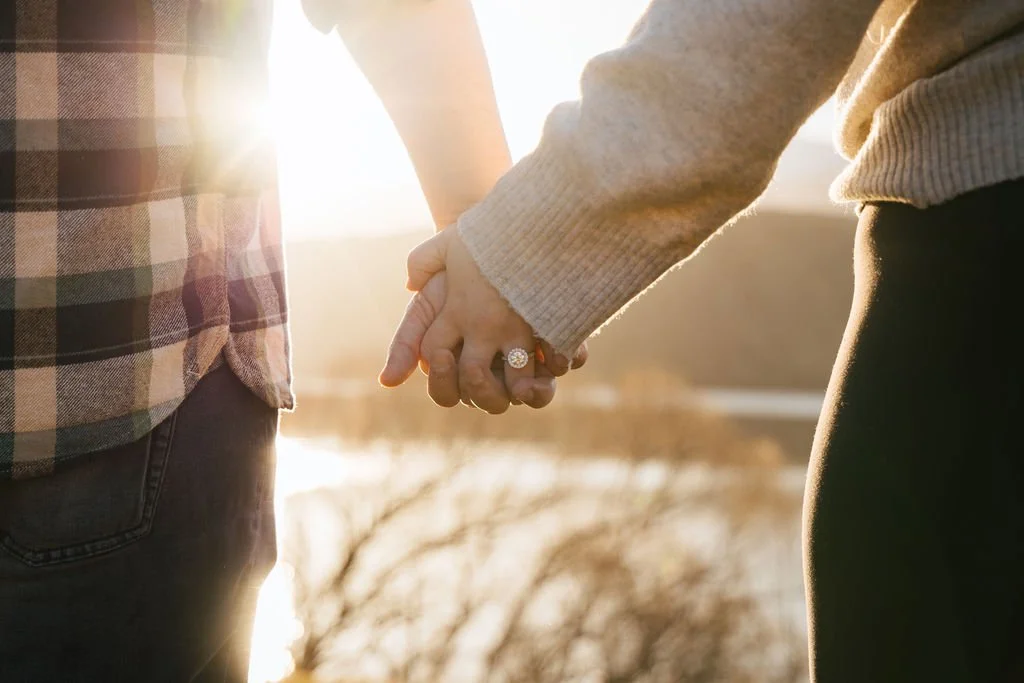 A couple holding hands outdoors during sunset, with the focus on their hands and an engagement ring on the woman.