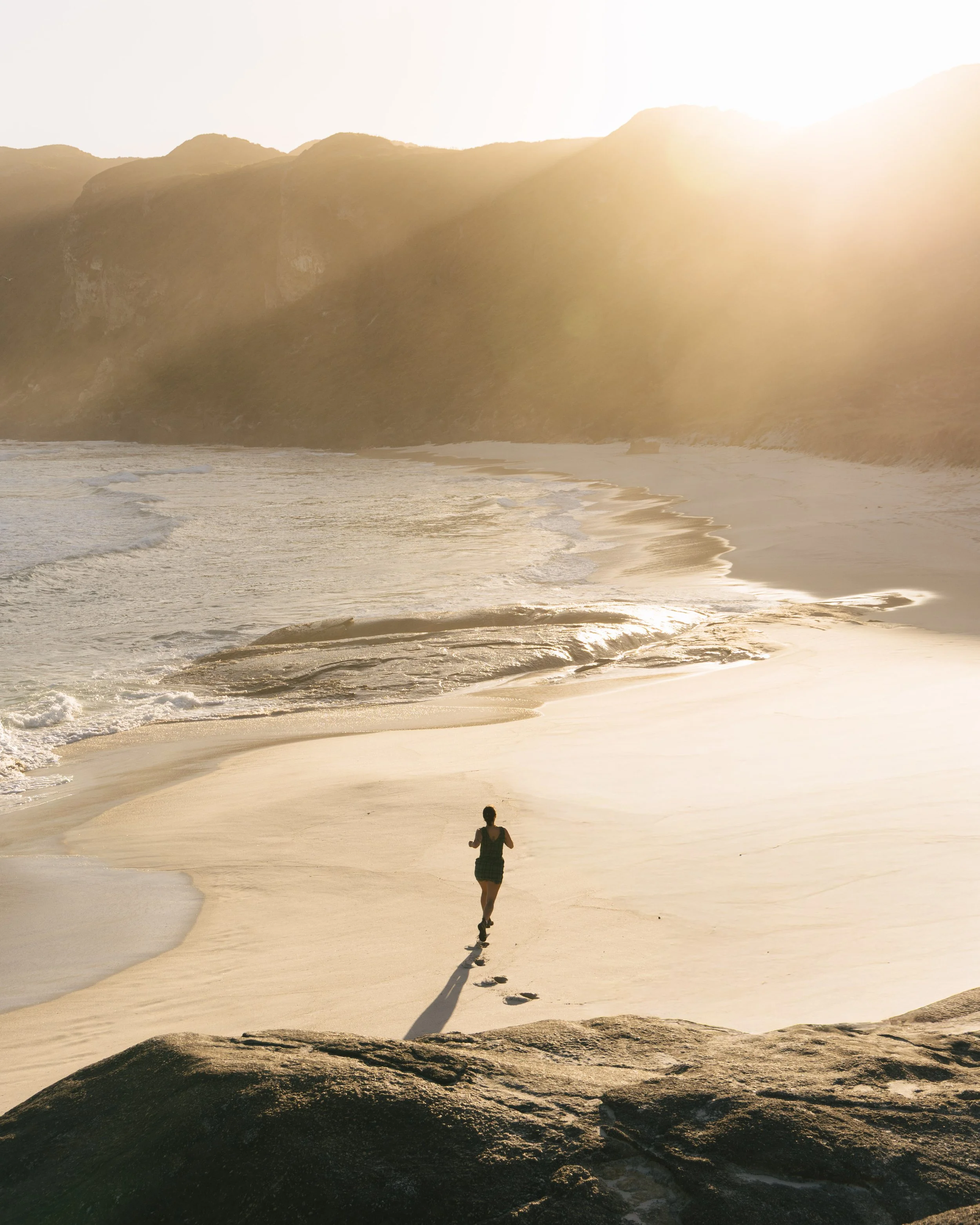 A person jogging on a sandy beach during sunset, with footprints behind them and hills in the background.