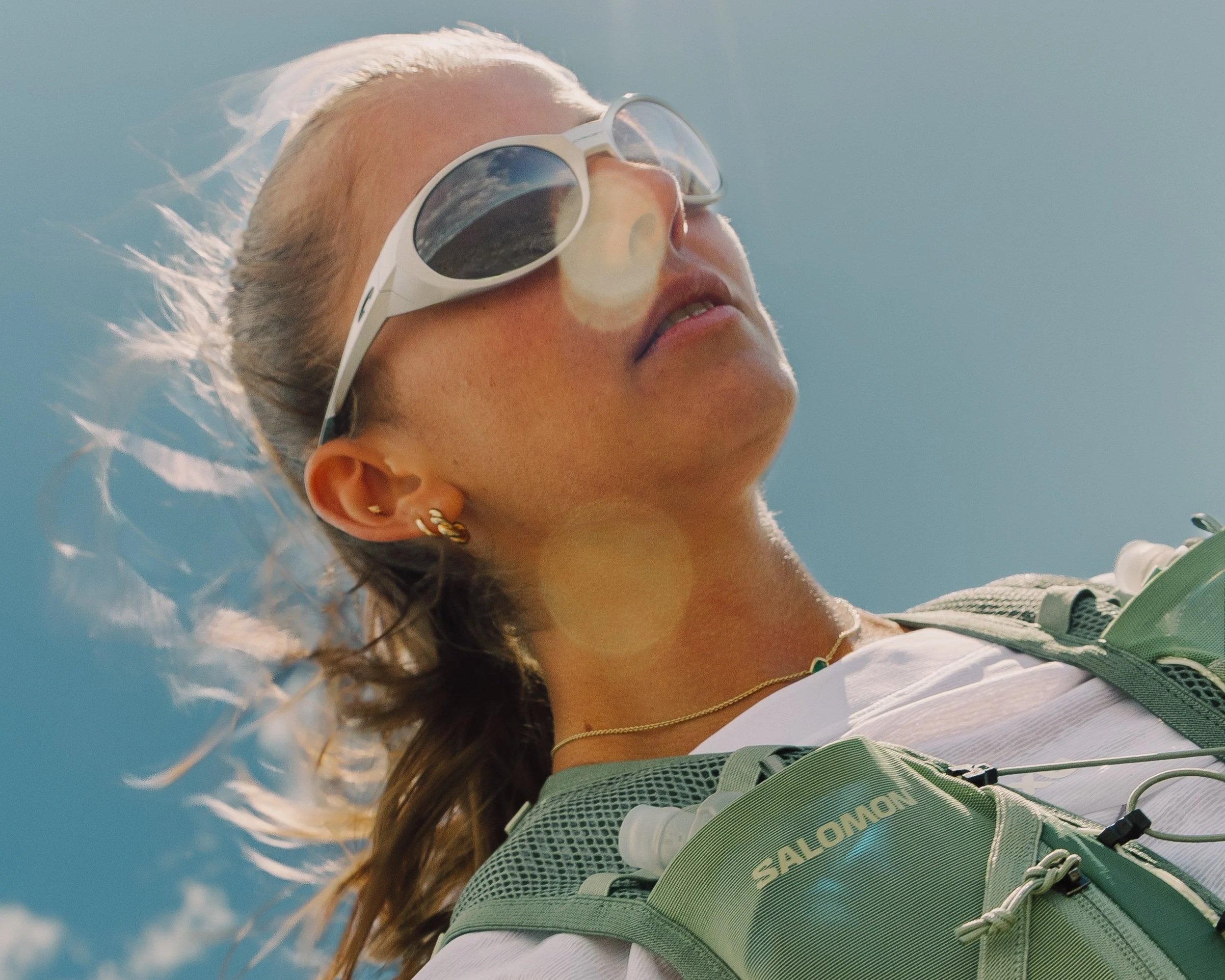A woman wearing white sunglasses, gold earrings, and a white shirt, carrying a green backpack, looking up against a blue sky with some clouds.
