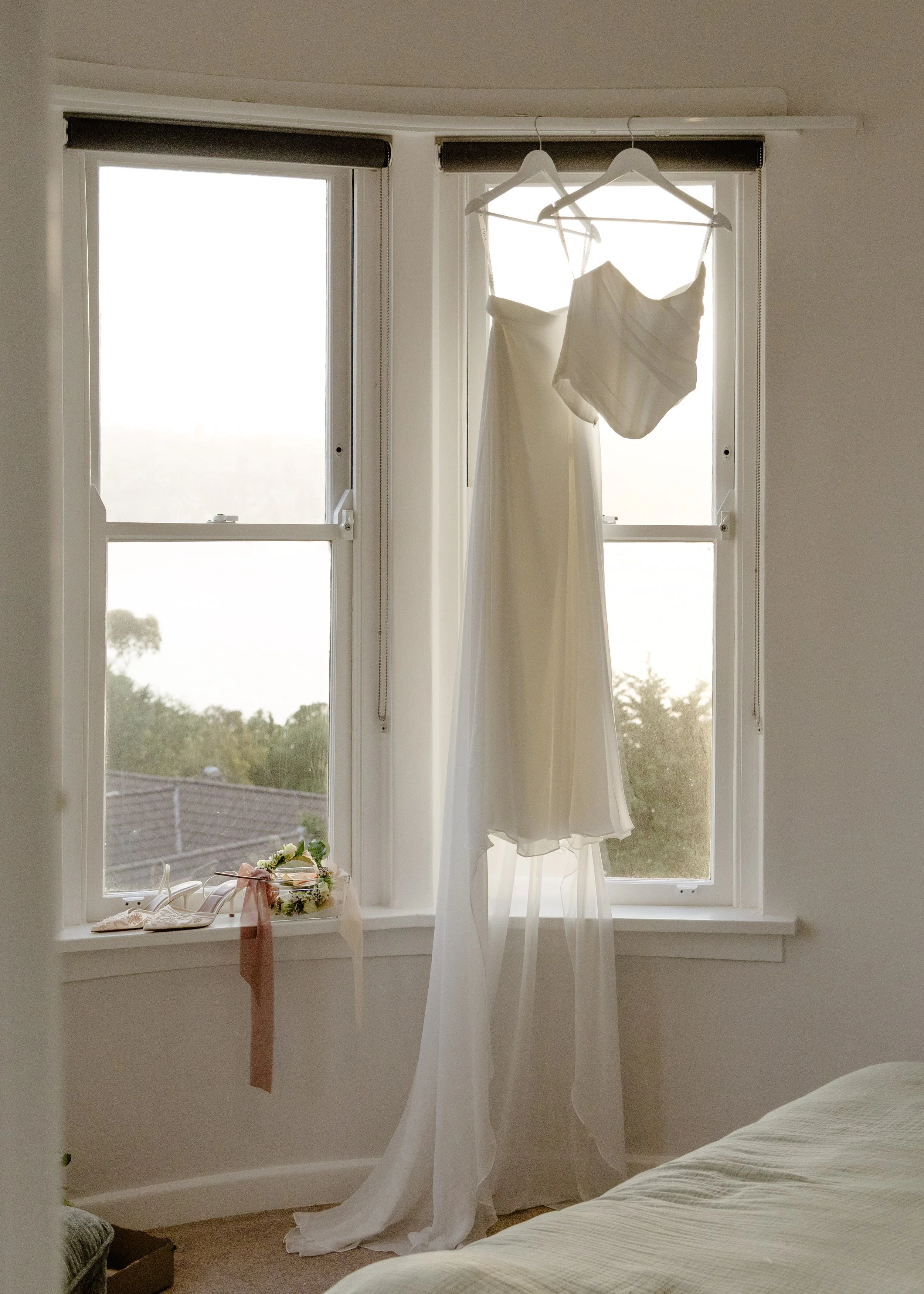 A wedding dress hanging in front of a window with sunlight streaming through, along with some wedding accessories on the windowsill.