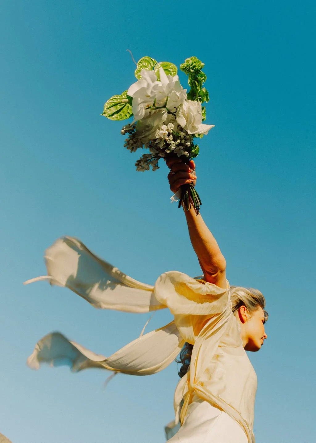 A woman in a flowing cream-colored dress holding a bouquet of white flowers and green leaves, raising it high against a clear blue sky.