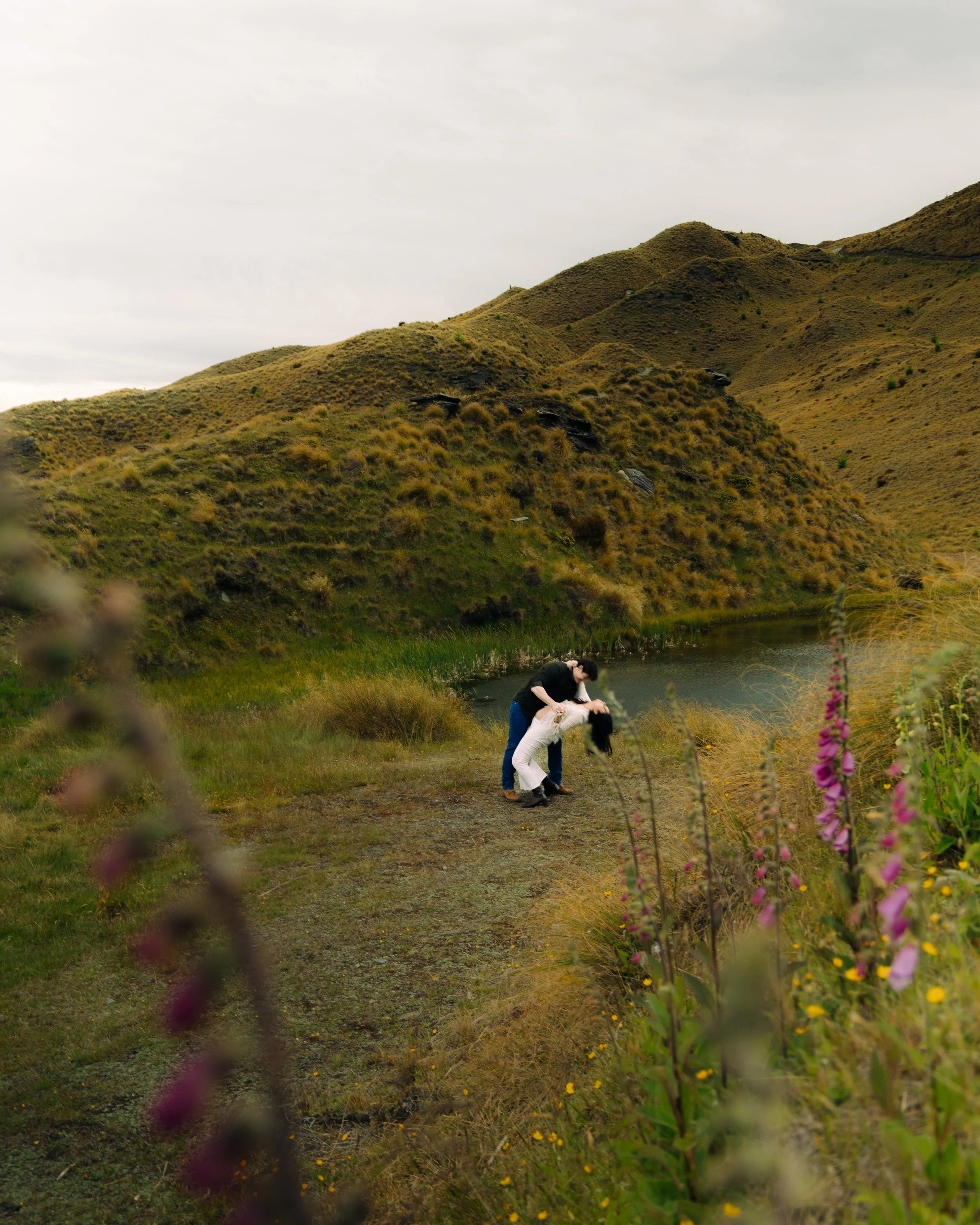A couple practicing acro yoga near a small pond in a hilly, rural landscape with wildflowers and grass, under a cloudy sky.