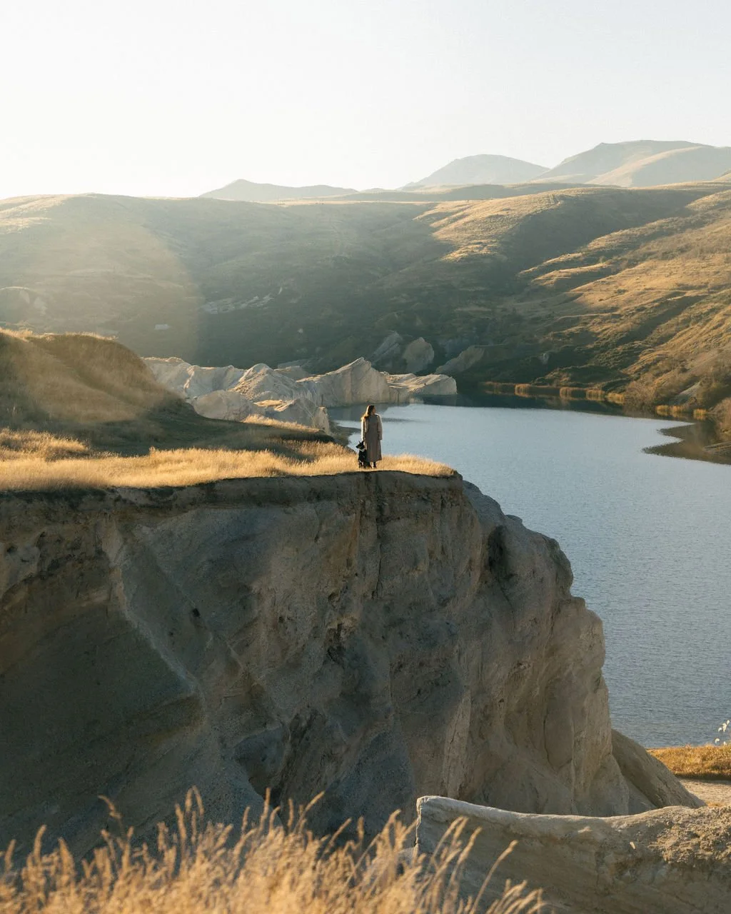 A person standing on a rocky cliff overlooking a lake with hills in the background during golden hour.