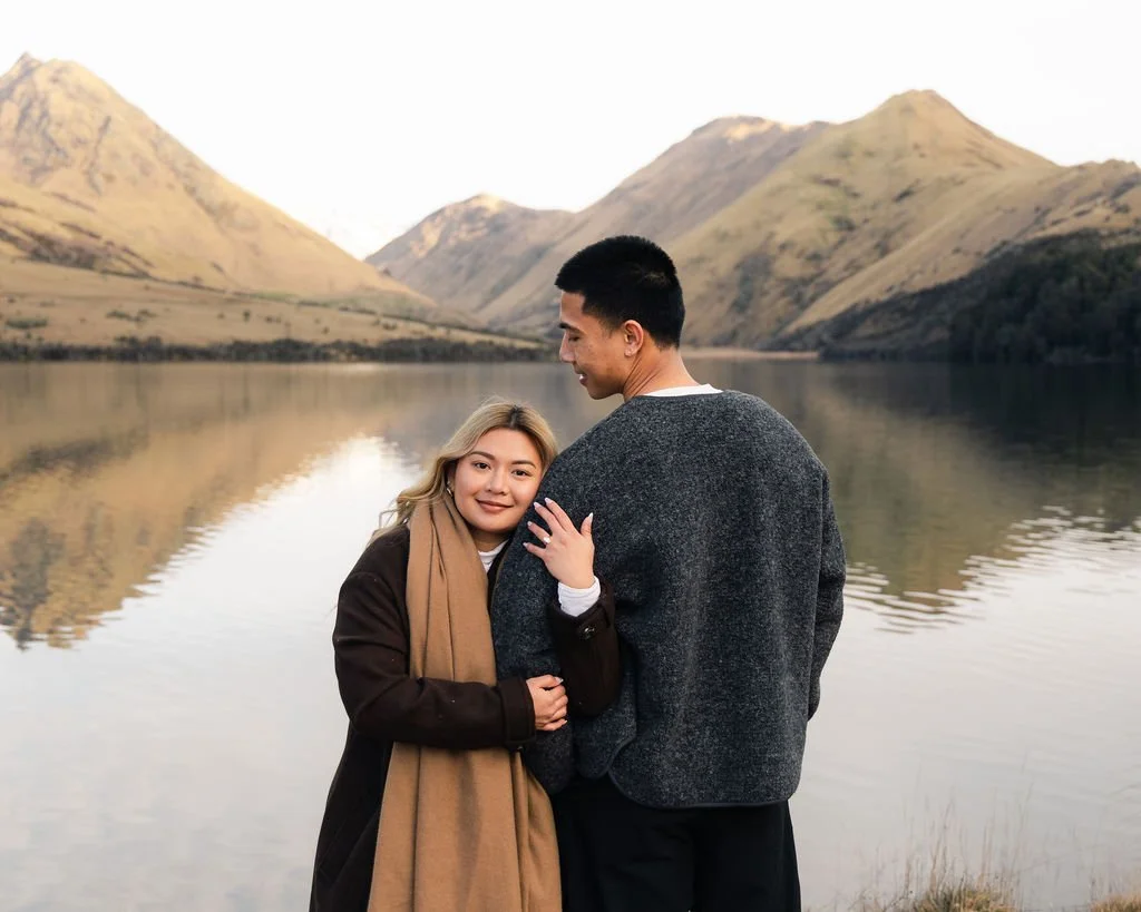 A young woman with blonde hair is hugging a tall young man with black hair, standing at a lakeside with mountains in the background. The woman is smiling and has her hand on the man's chest, while the man is looking down at her. They are dressed warmly, indicating cool weather.