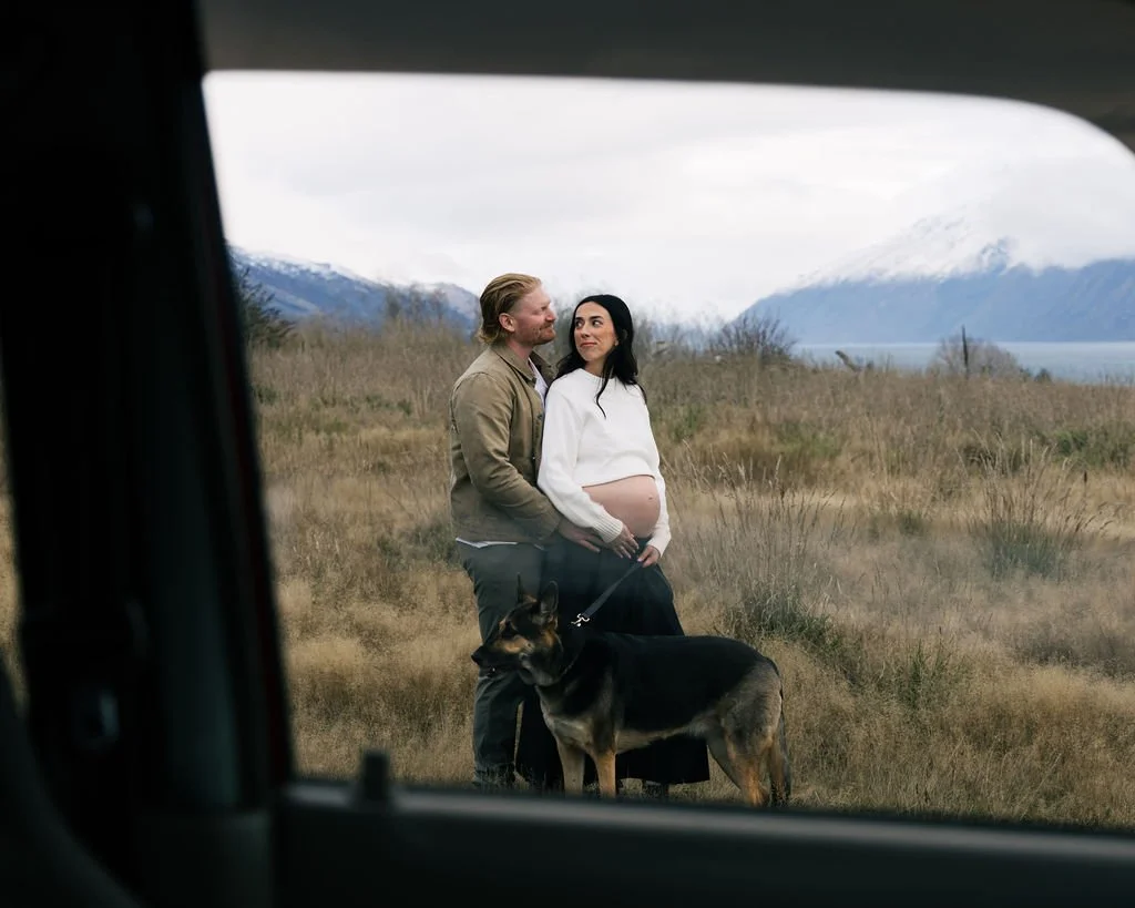 A couple and their dog standing in a field, with snow-capped mountains and a body of water in the background, viewed from inside a vehicle.