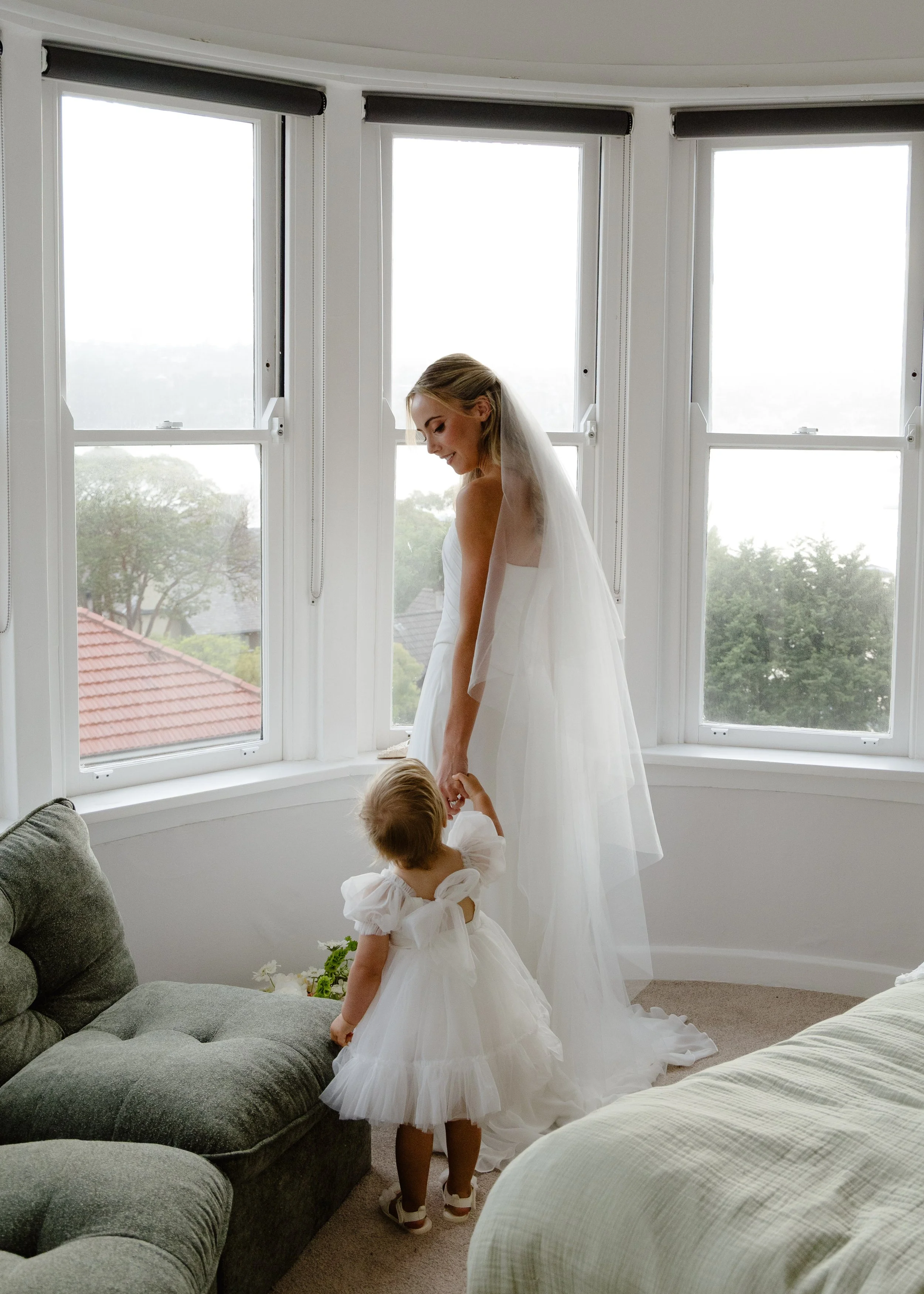 A bride in a white wedding dress and veil holding hands with a young girl in a white dress with puffed sleeves, inside a room with large bay windows overlooking some trees and rooftops.