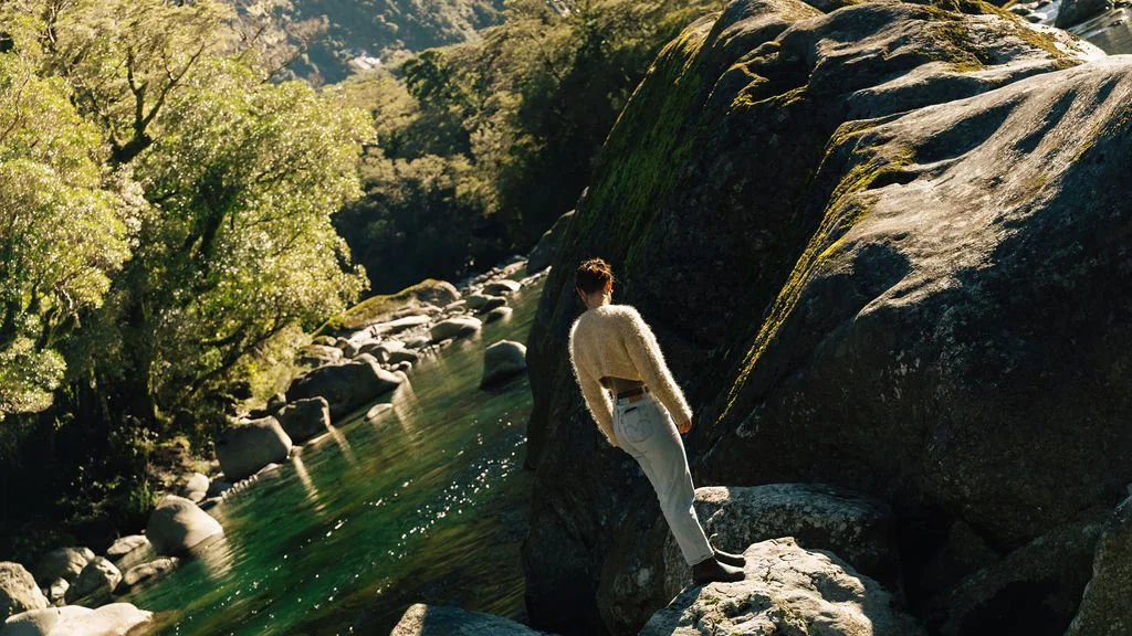 Person leaning on a large moss-covered boulder beside a river in a forested area.
