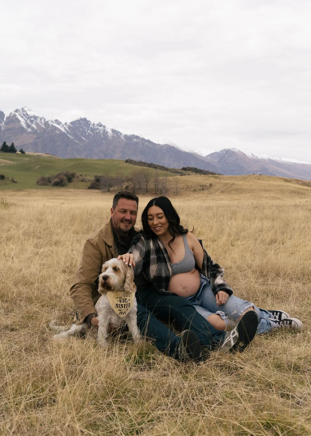 A couple sitting on a grassy field with a dog, mountain range in the background, cloudy sky.