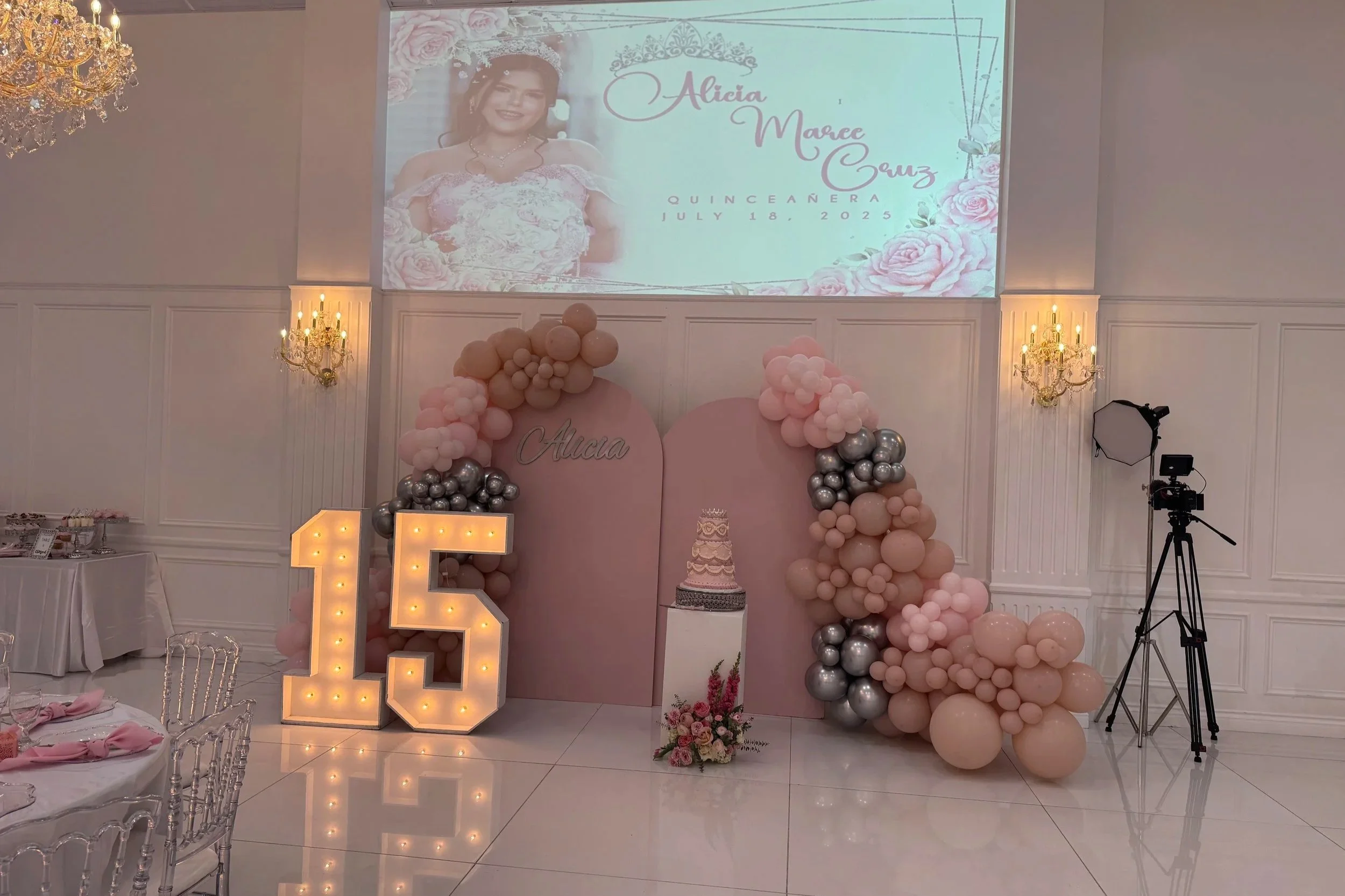 Sweet 15 celebration decoration with a pink and silver balloon arch, a large illuminated number 15, a pink backdrop with a cake on a stand, and a projection screen displaying a photo of a girl in a wedding dress with text about her event on July 18, 2025.