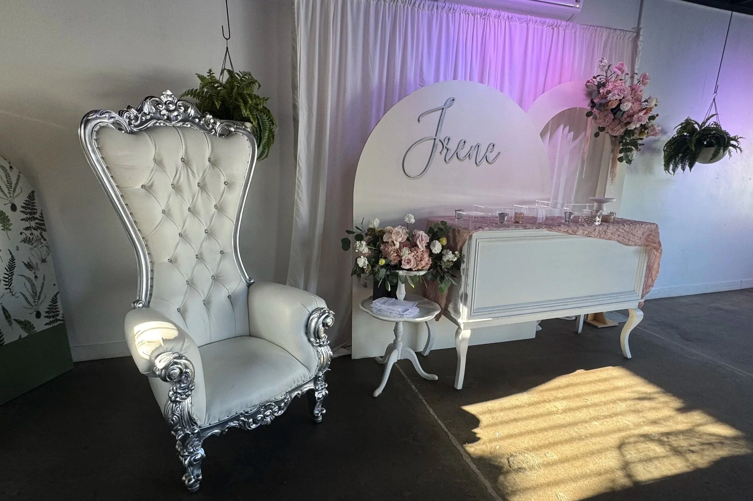 Elegant white throne chair with silver accents, floral arrangement on a small table, and a white backdrop with pink and white flowers, with a sign reading 'Jrene' in cursive, set up for a celebration or event.