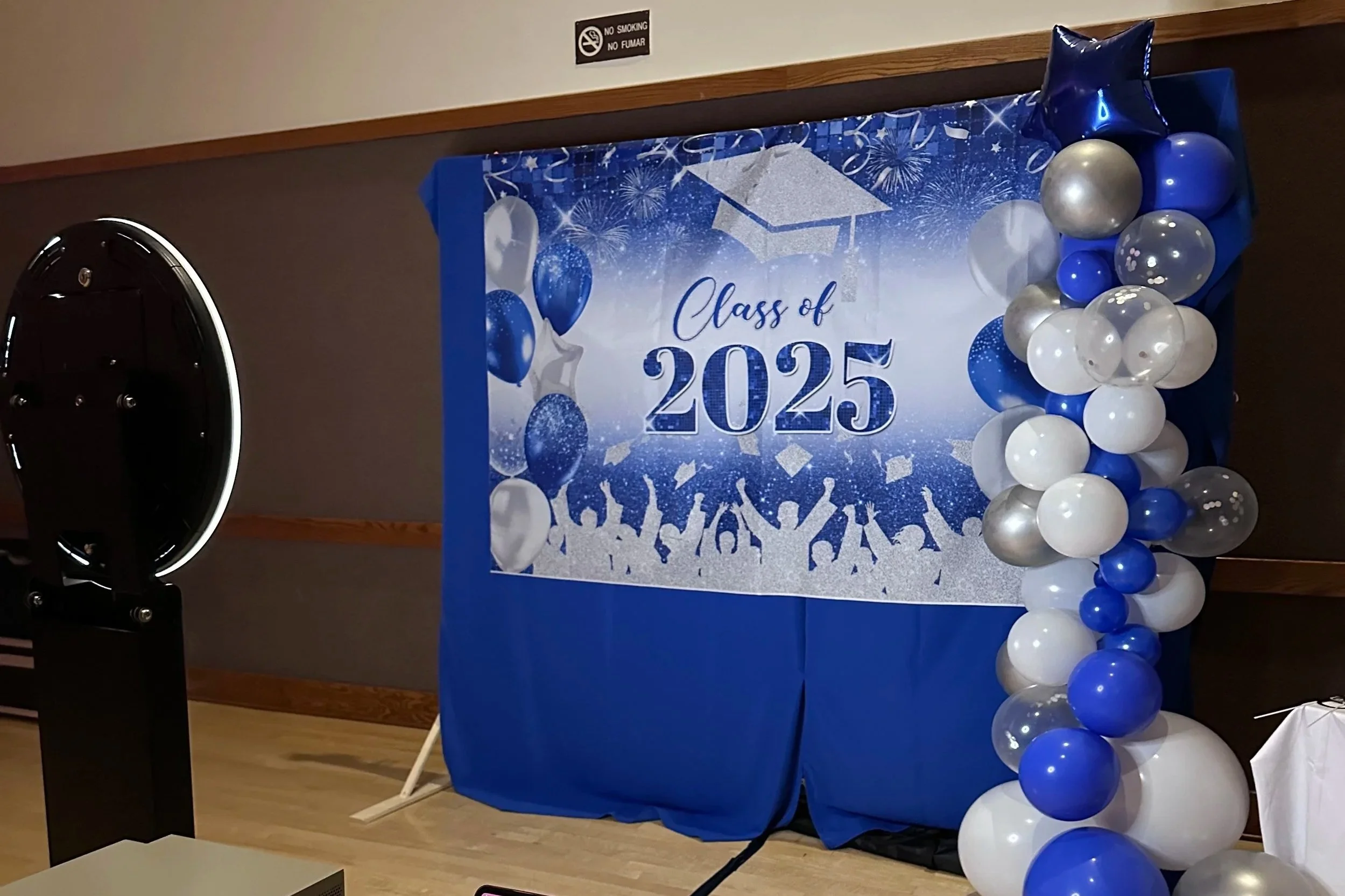 Graduation celebration backdrop with balloons for the class of 2025, featuring a blue and white color theme and silhouettes of cheering graduates.