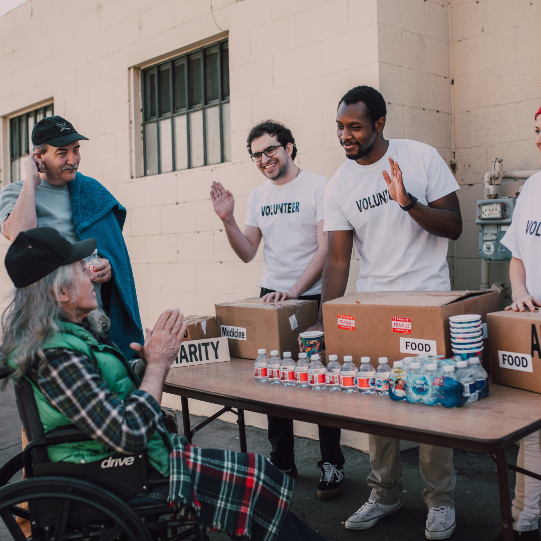 A group of volunteers handing out food and bottled water to an elderly male in a wheelchair during a charity event outside a building.