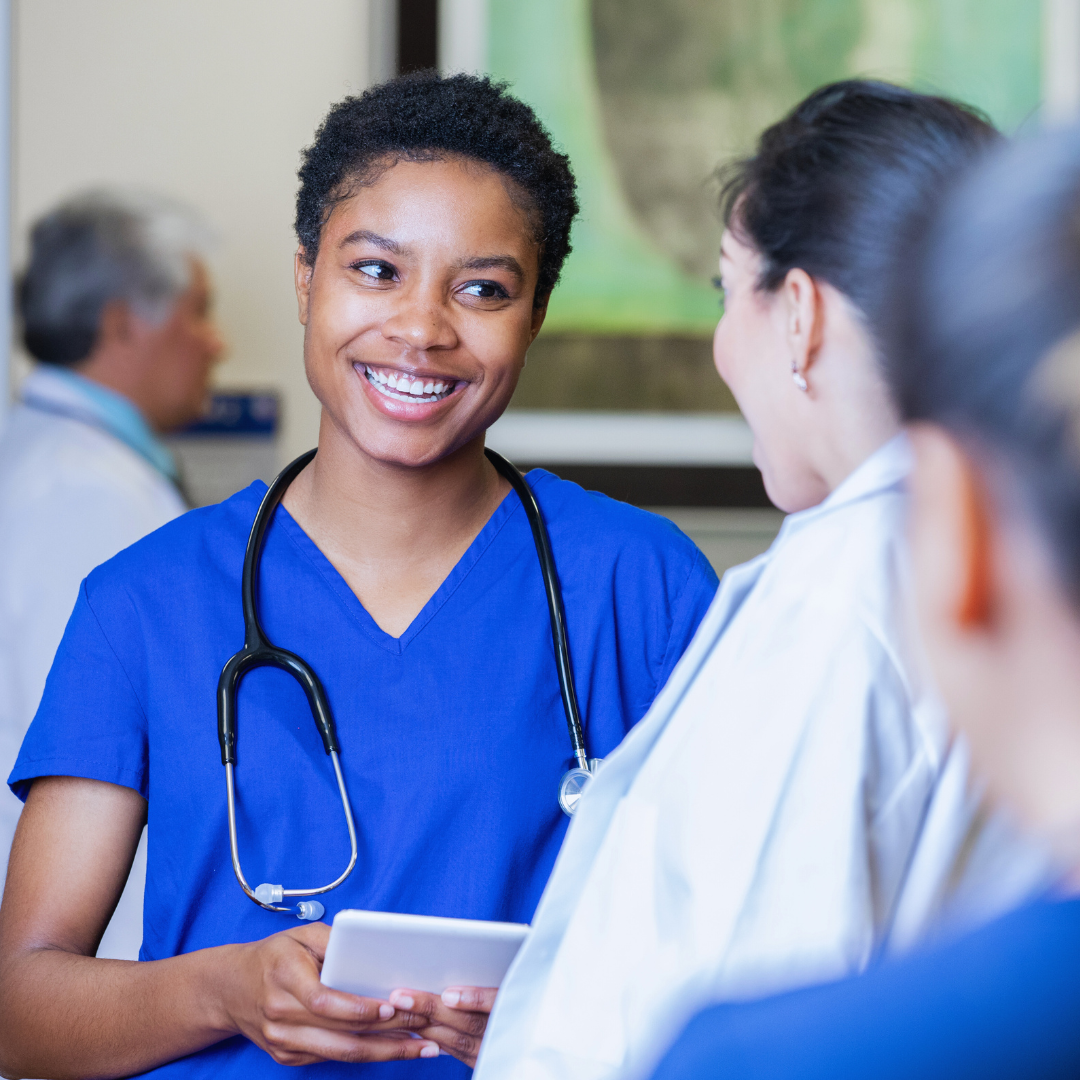 A smiling female nurse with a stethoscope around her neck talking to a colleague in a healthcare setting.
