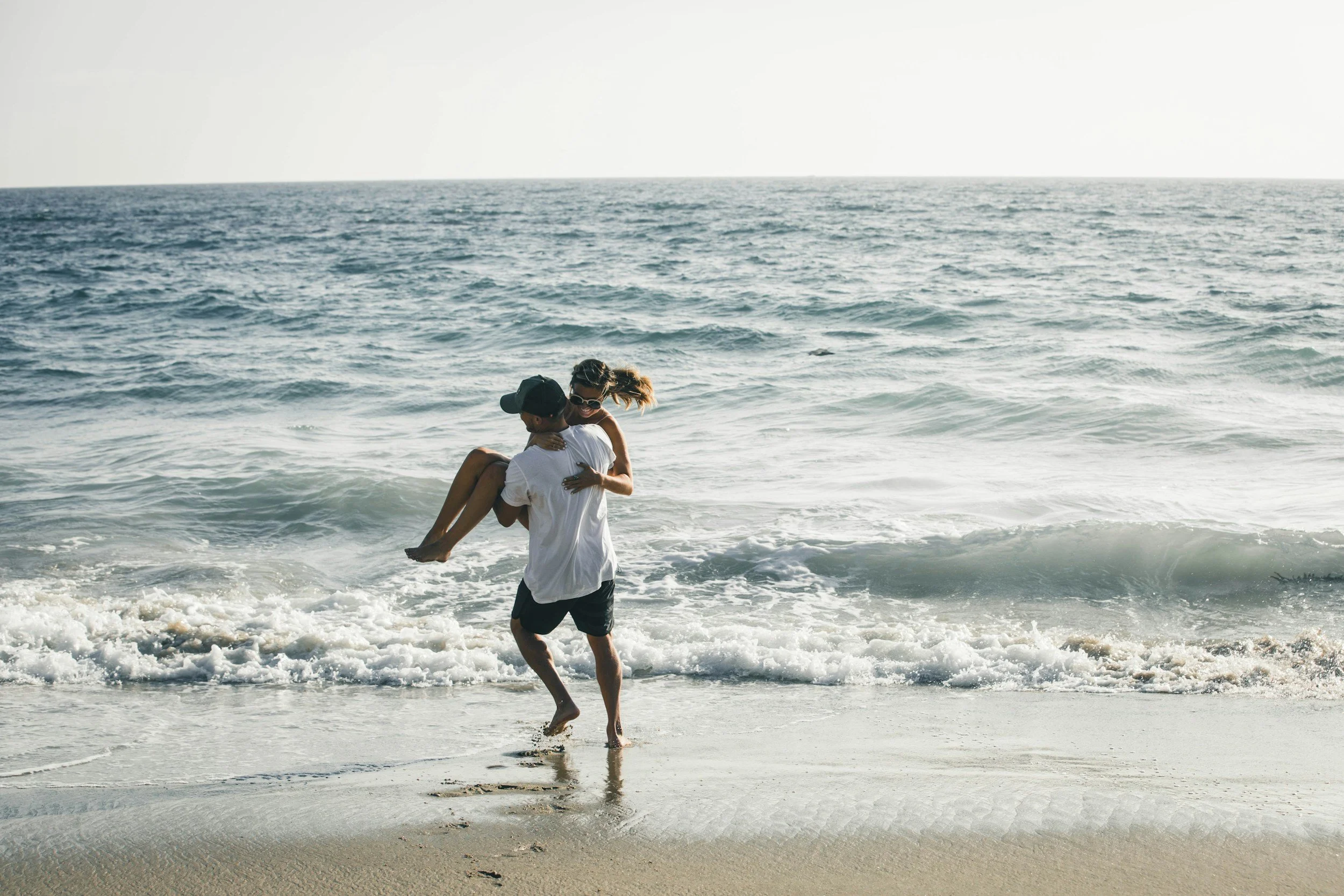 A man carrying a woman in his arms walking along the beach at the shoreline with waves crashing on the sand and ocean in the background.