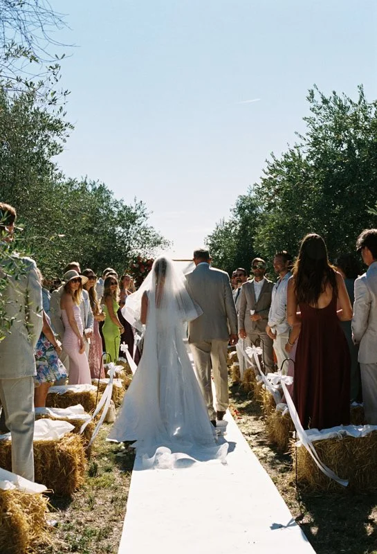 The bride walks with her father between bales of hay and olive trees to the outdoor wedding ceremony; analogue takes photos