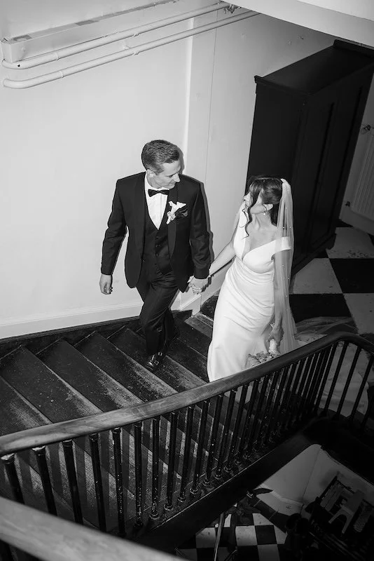 The bride and groom walk up a staircase hand in hand, looking at each other.