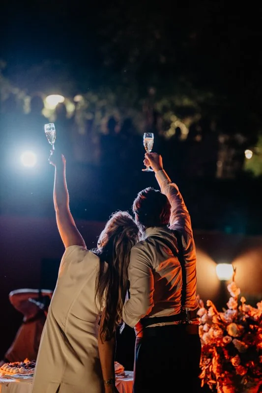 The bride and groom toast each other in the evening with raised champagne glasses, photographed from behind
