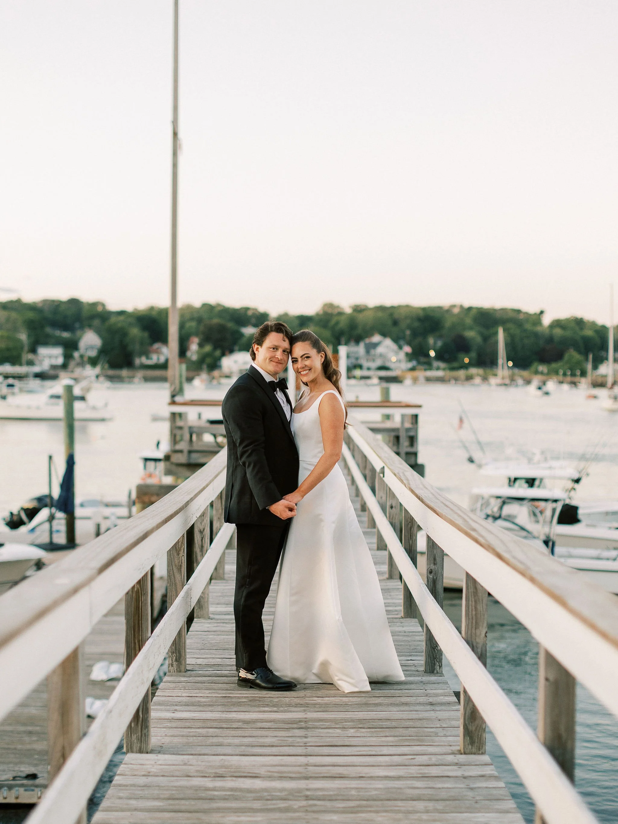 A newly married couple in formal wedding attire standing on a wooden dock by the water, holding hands and smiling, with boats and houses in the background.