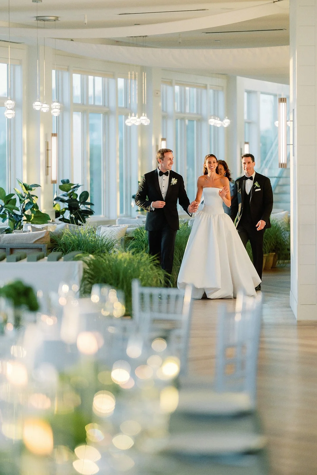 A bride and two grooms walking in a bright, modern venue decorated with green plants and soft lighting.