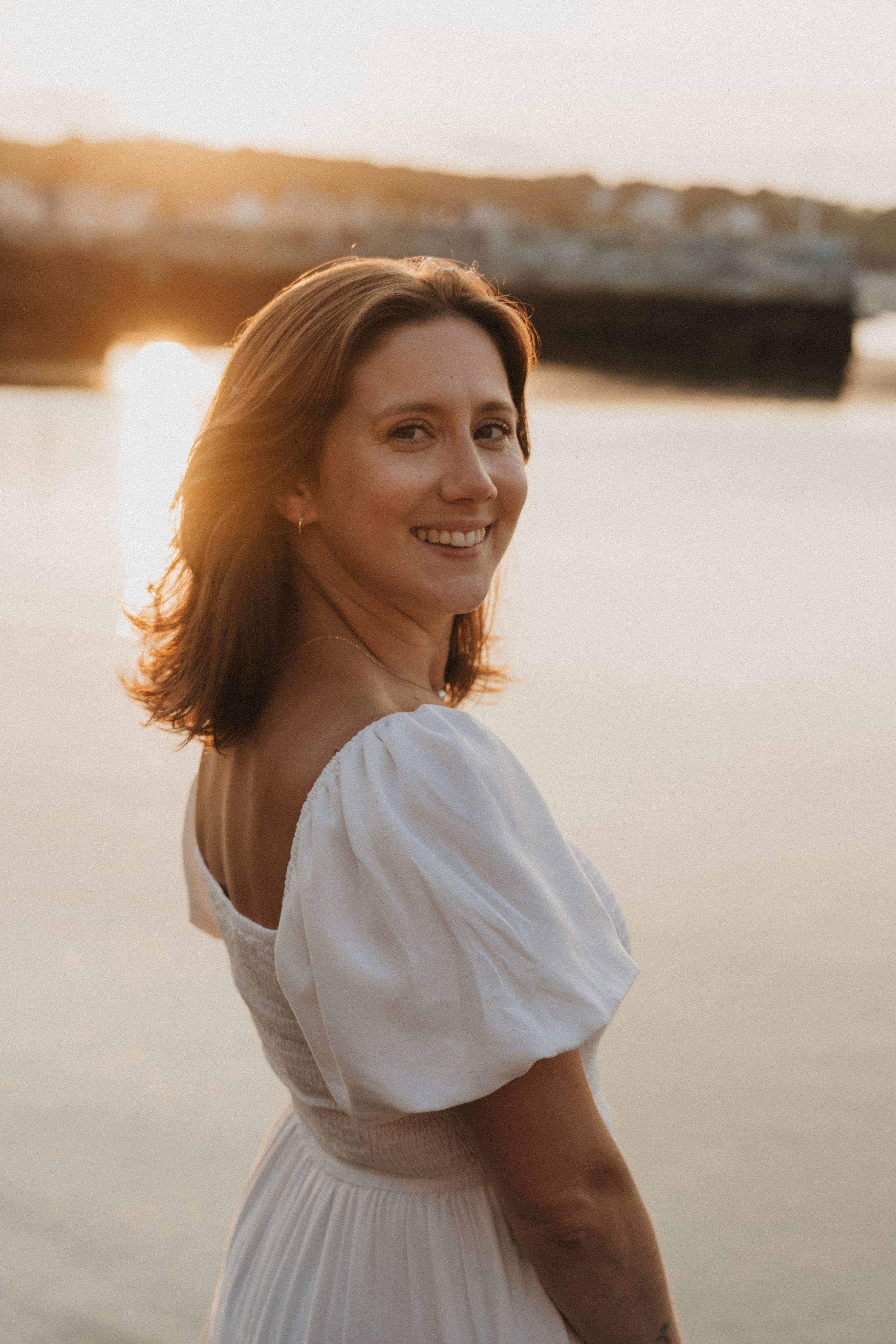 A woman with shoulder-length brown hair smiling at the camera, wearing a white dress with puffed sleeves, standing near water with a yacht and sunset in the background.
