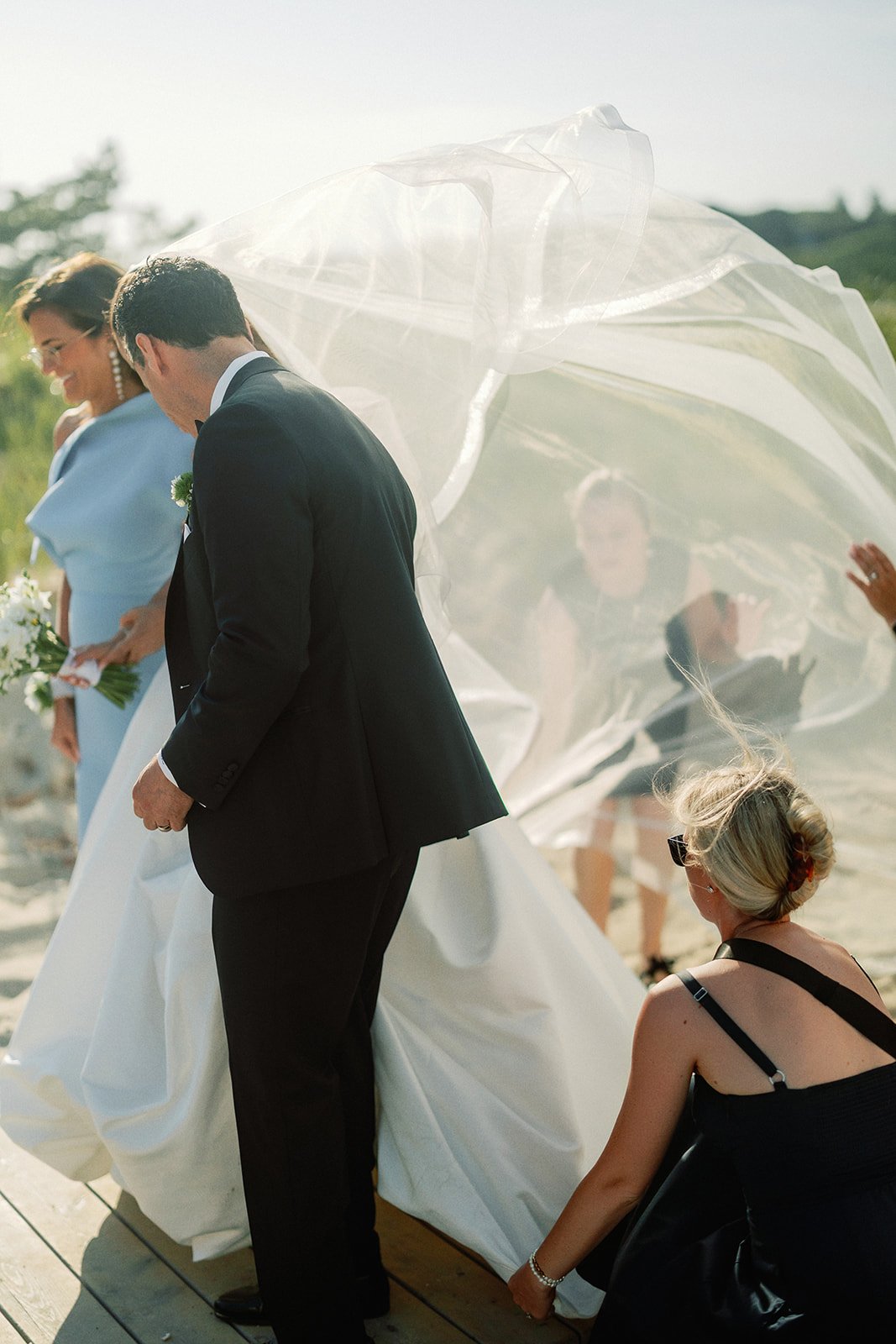 A group of people, including a woman in a light blue dress holding a bouquet and a man in a black tuxedo, are on a beach during a wedding ceremony. A woman dressed in black is crouching on the sand, and another woman in the background is smiling while adjusting her dress. A large, flowing white fabric or veil is blowing in the breeze.