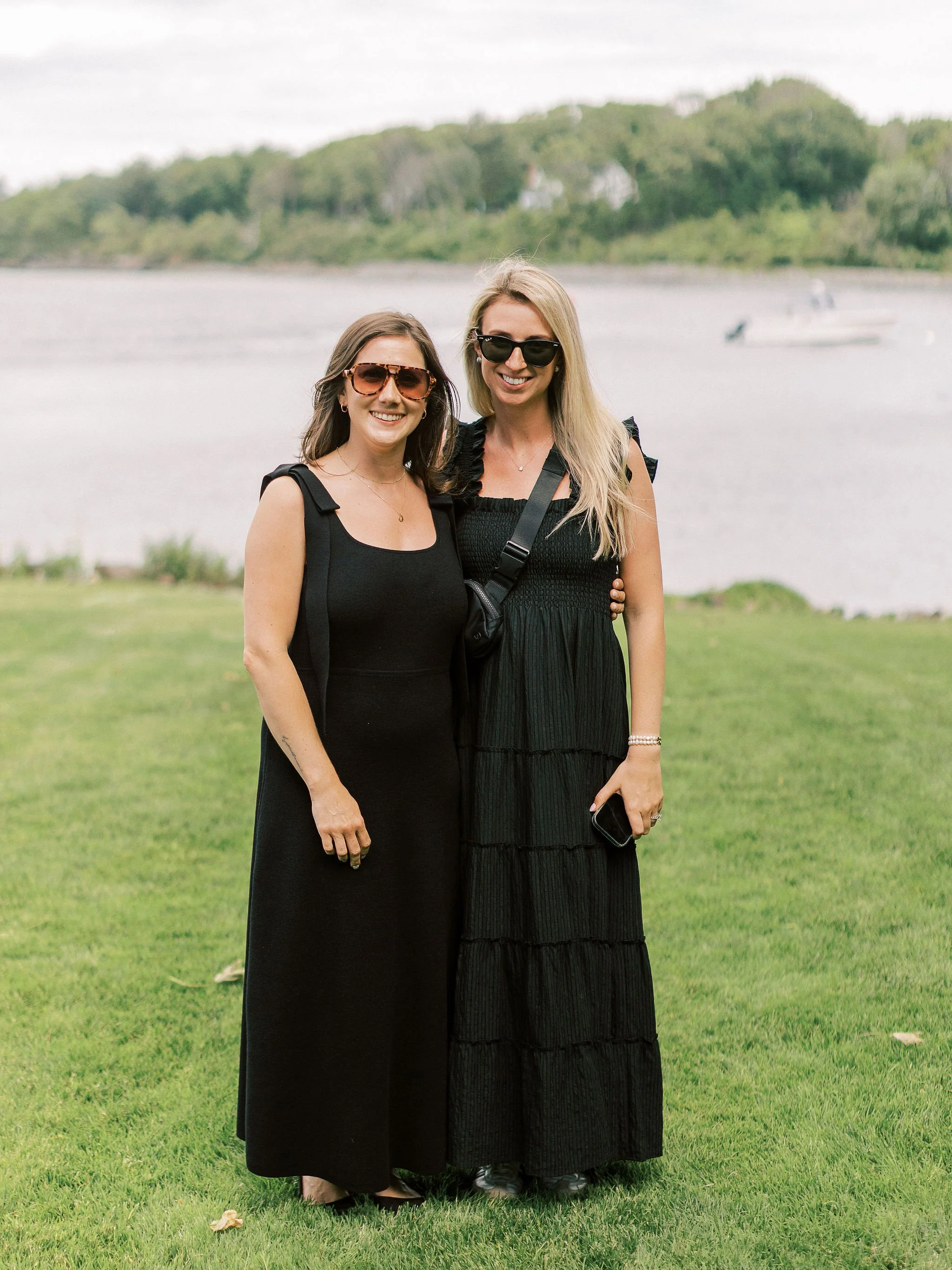 Two women standing on grass near a body of water with boats and trees in the background, both wearing black dresses and sunglasses, smiling at the camera.