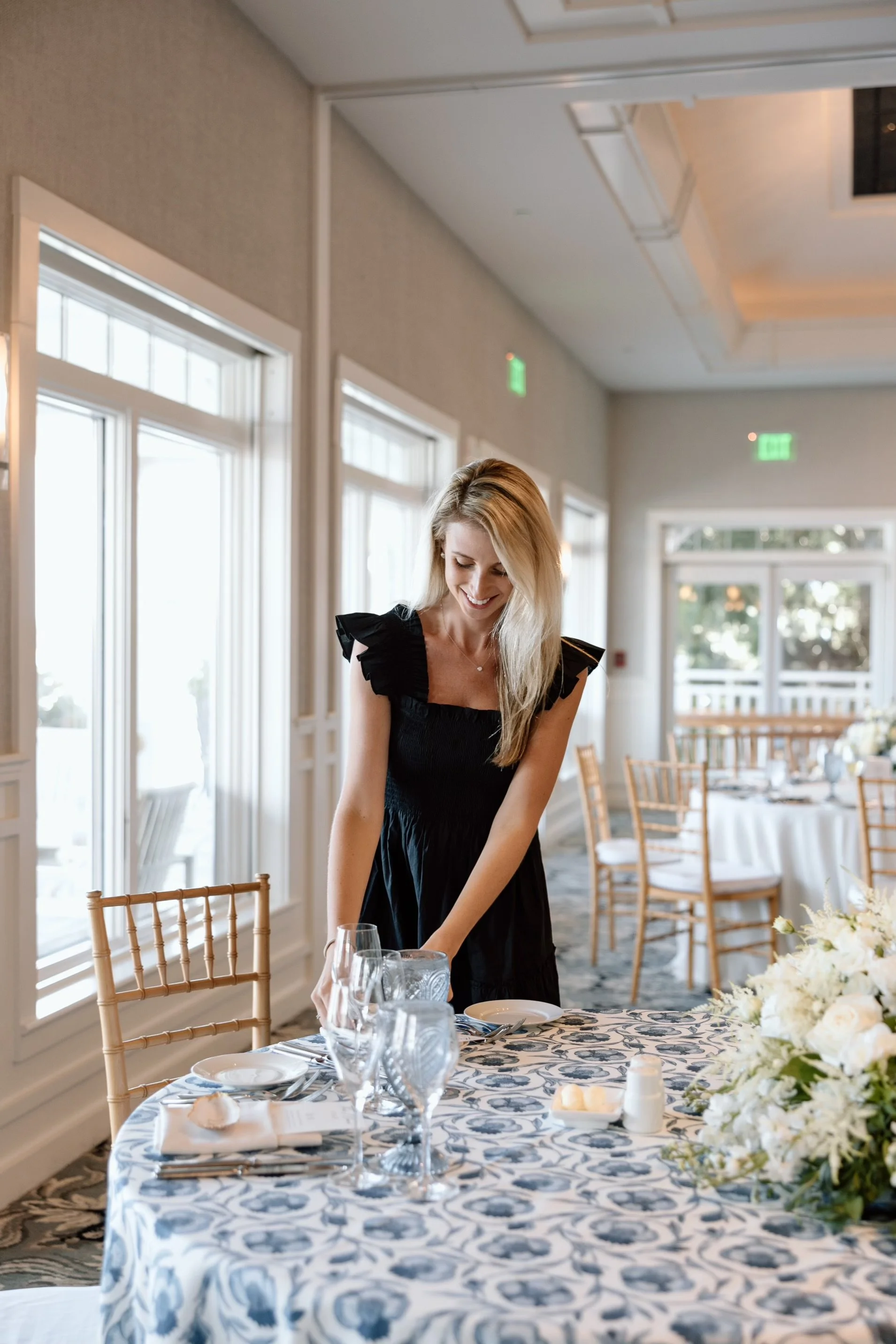A woman in a black dress setting a table in a bright, elegantly decorated dining room with large windows, white walls, and a floral tablecloth.