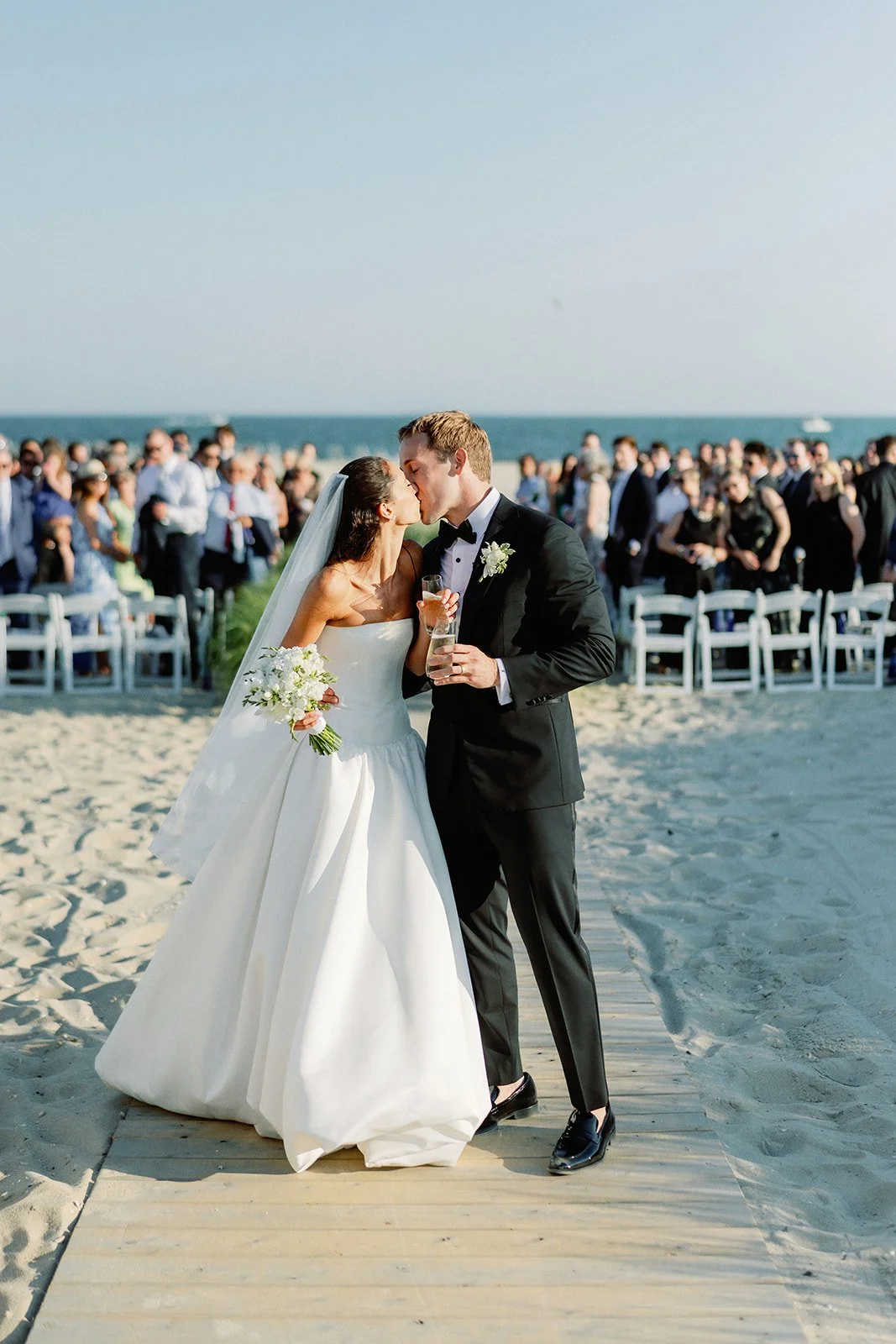 Bride and groom kiss at their beach wedding, surrounded by guests, on a sandy path with the ocean in the background.