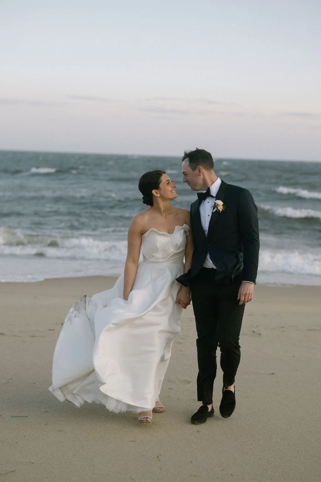 A bride and groom on the beach, holding hands and smiling at each other, with the ocean waves in the background during sunset or dusk.