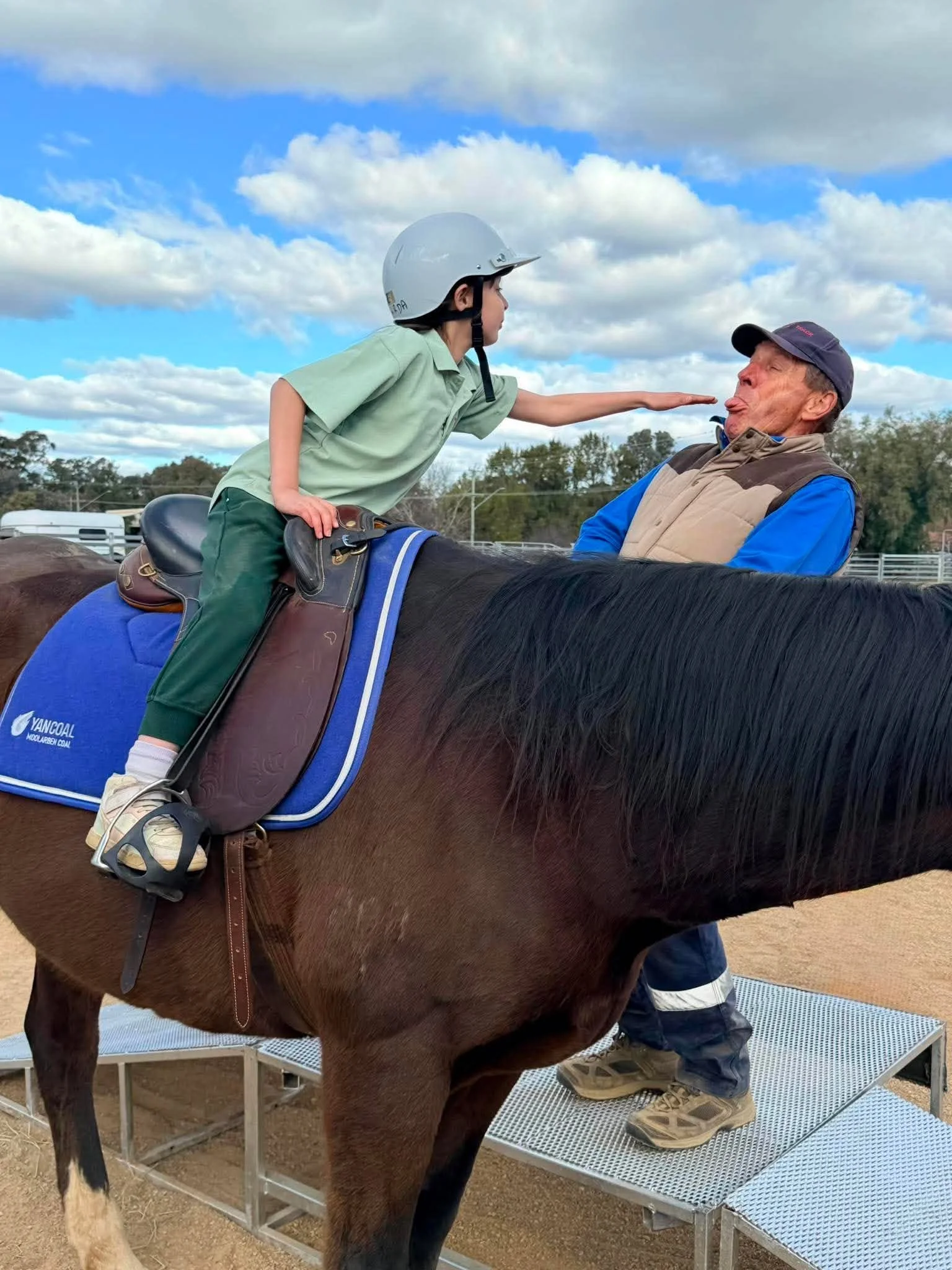 A young girl in a helmet riding a horse with an elderly man in a cap sitting on the horse, making a funny face with his tongue out, outdoors under a partly cloudy sky.