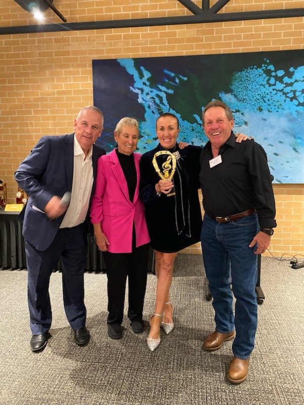 Four people standing together at an award ceremony, with three men and one woman holding a trophy. The setting is indoors with a brick wall and an abstract blue painting in the background.