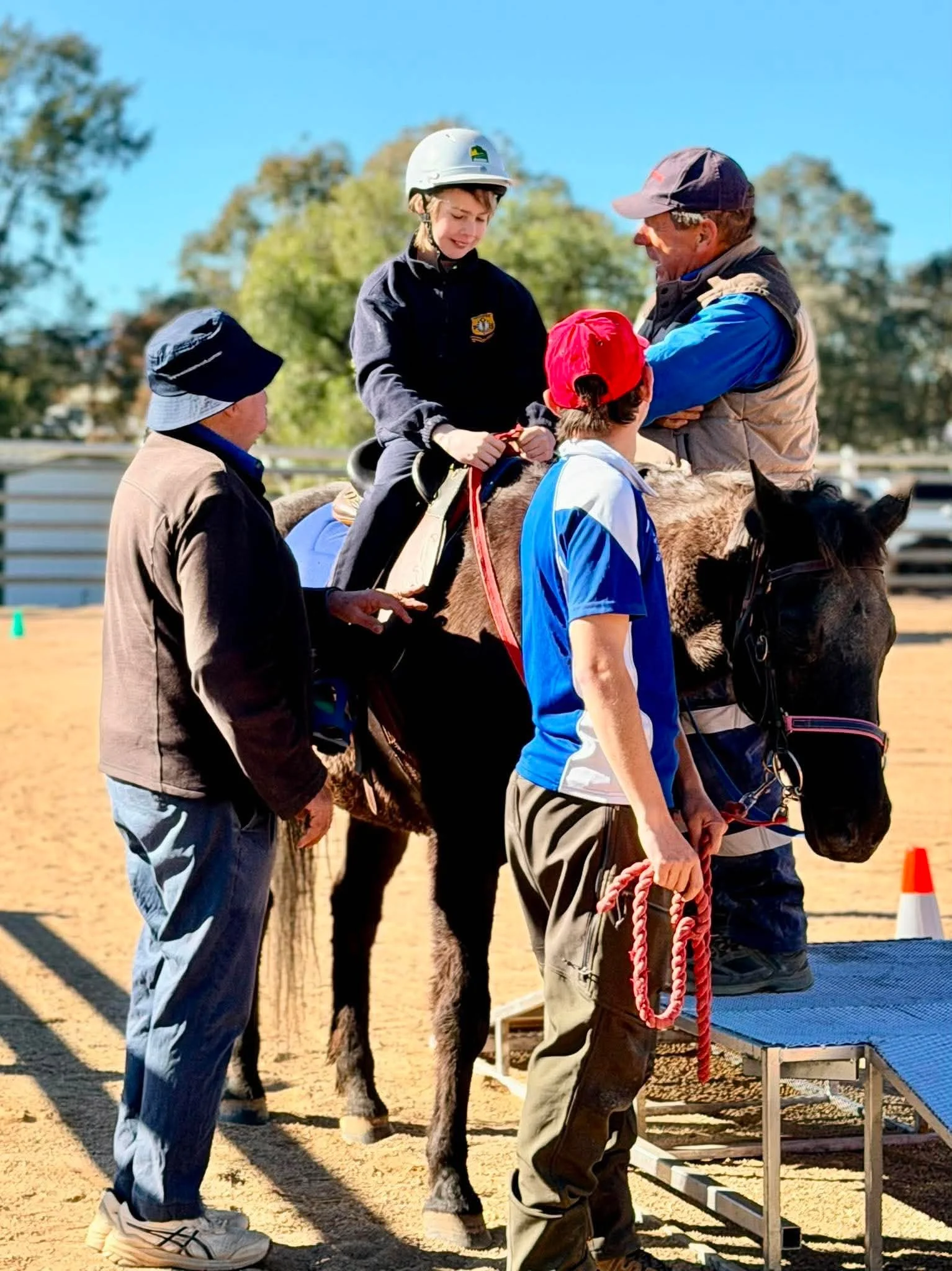 Young girl riding a horse with adults assisting during an outdoor event on a sunny day.