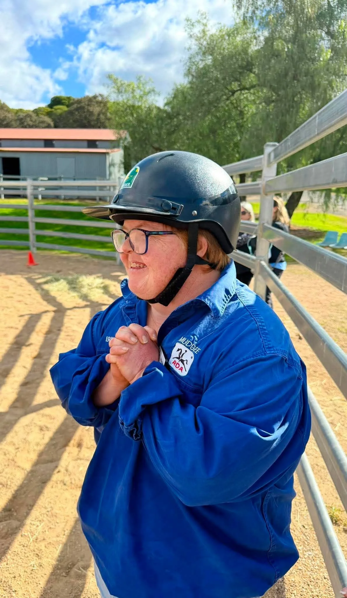 A woman wearing a black helmet, glasses, and a blue jacket stands outdoors with her hands clasped, smiling. Background includes trees, a metal fence, and a building under a partly cloudy sky.