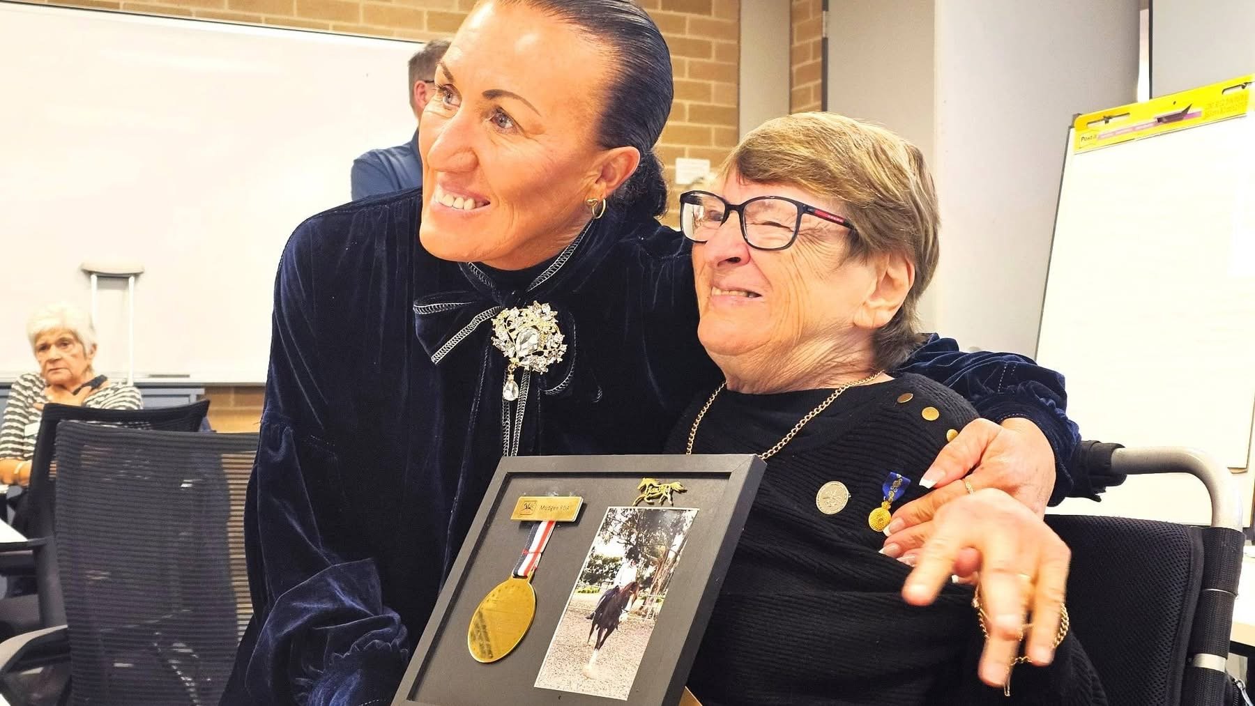 A woman in a dark dress and glasses is sitting in a wheelchair, holding a framed photo and medals. A woman with dark hair and a bolo tie is hugging her and smiling. In the background, another woman with white hair is sitting and watching.