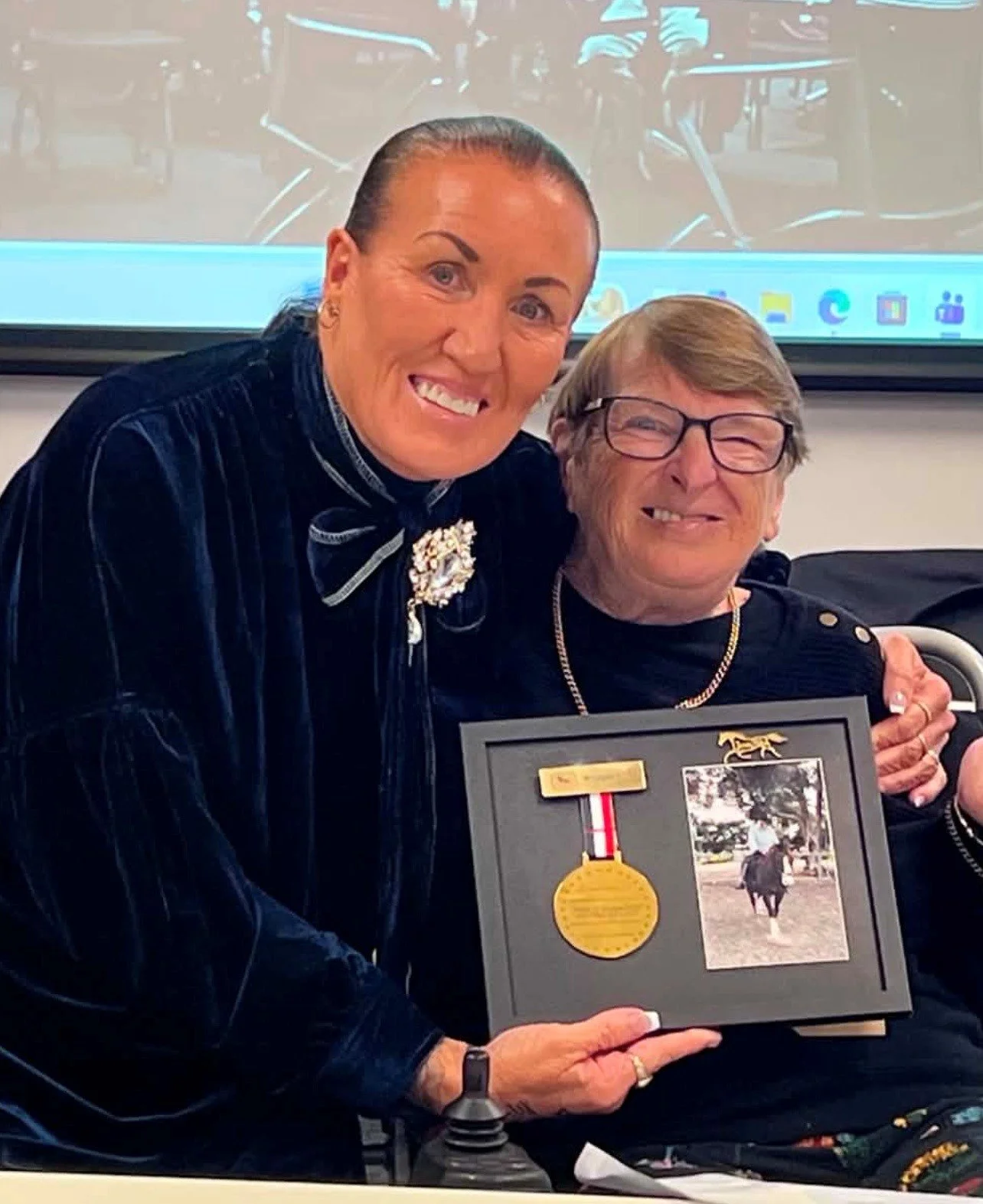 Two women smiling, one holding a framed photo and medal, with a presentation screen in the background.