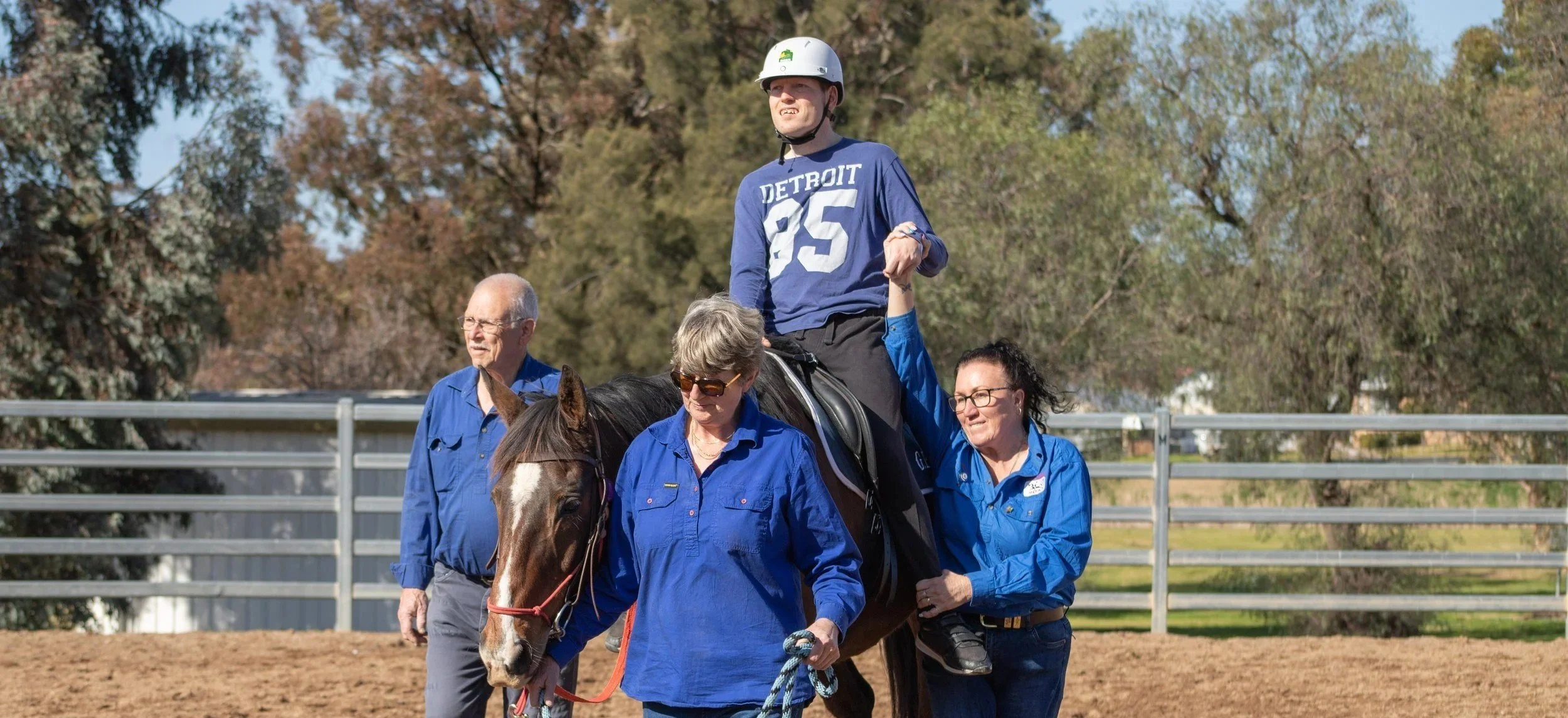 A young man  riding a horse, accompanied by several adults helping guide the horse. There are trees and a fence in the background on a sunny day.