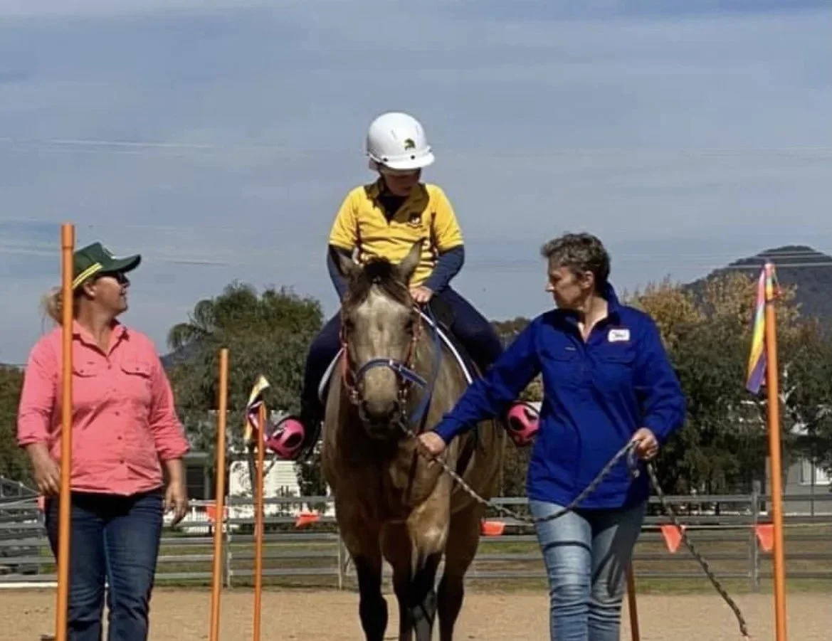 A woman in a pink shirt and green cap walking next to a horse with a rider in a yellow shirt and white helmet, while another woman in a blue jacket holds the horse's reins in an outdoor riding arena.