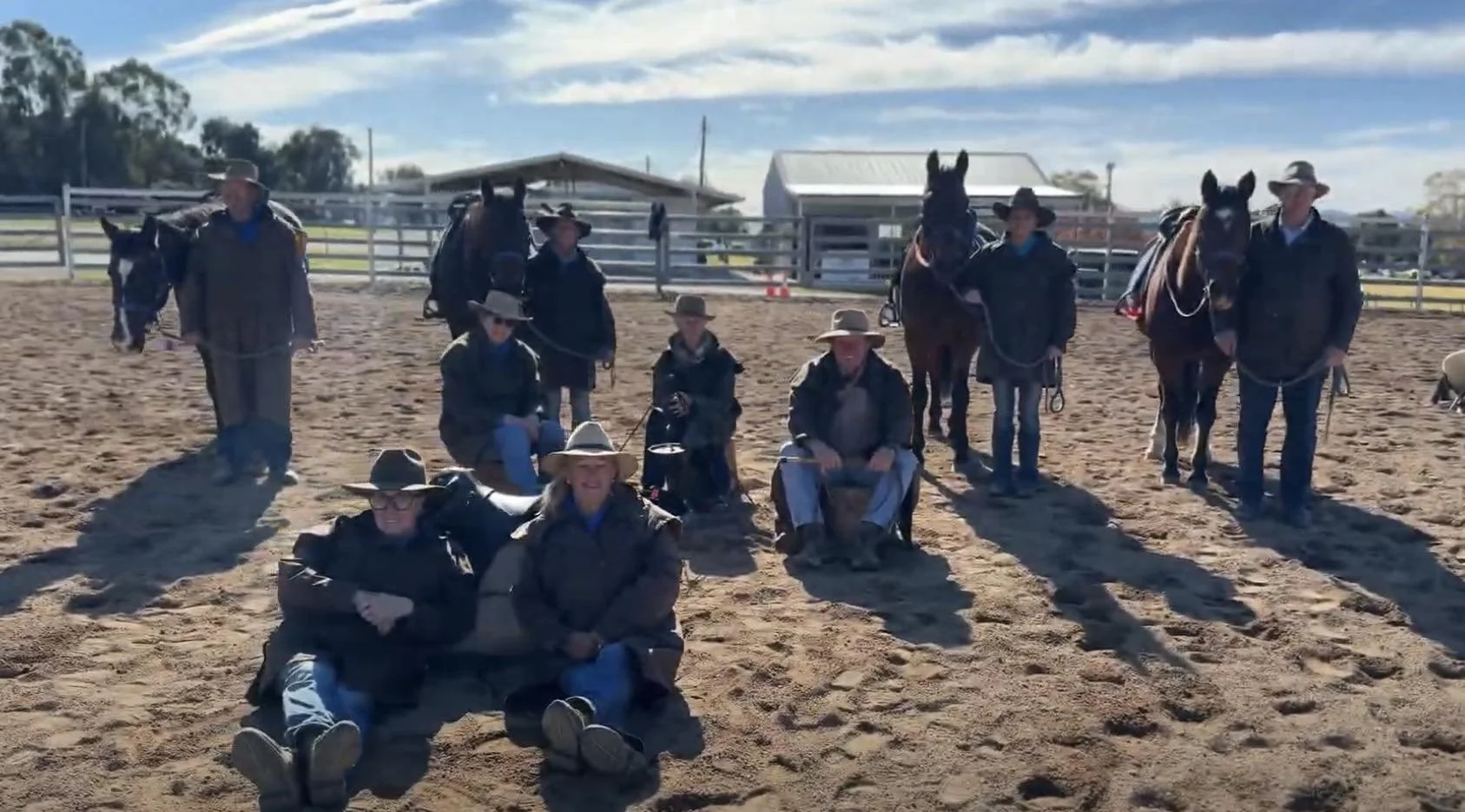 Group of people with hats and jackets sitting and standing with horses in a dirt riding arena, with fences and buildings in the background on a sunny day.