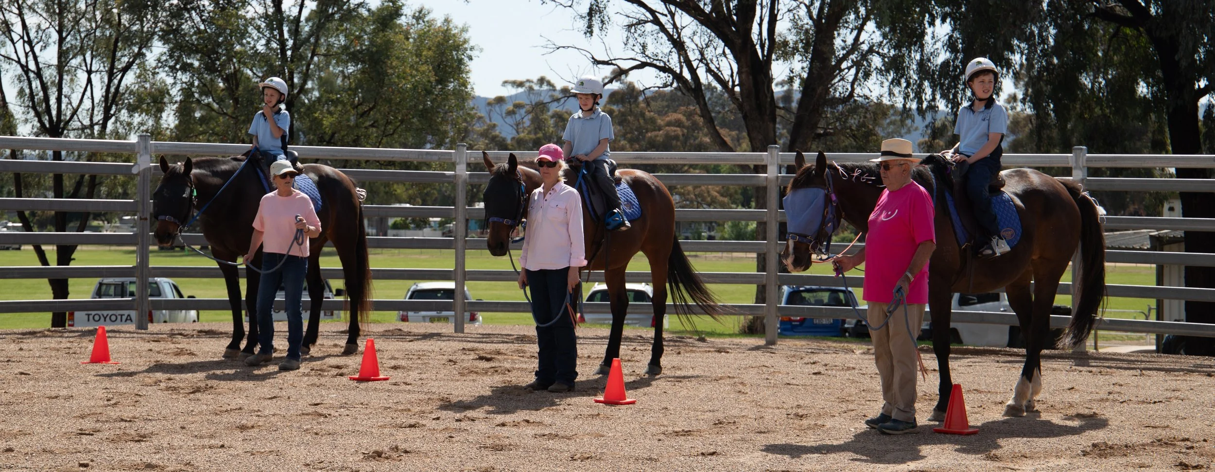 Children riding horses in an outdoor riding arena with instructors guiding them, orange traffic cones marking the course, and trees in the background.