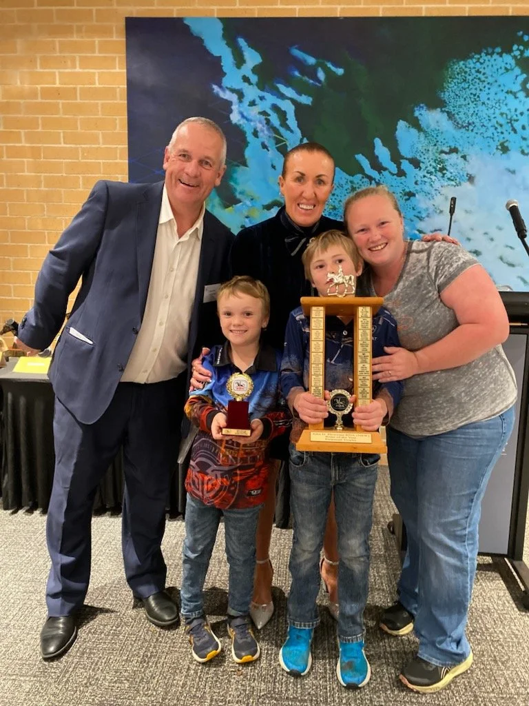 A group of five people, including two children, posing happily indoors. The children are holding trophies. The group includes three adults, one man in a blue suit and two women, one in black and the other in gray. A large abstract blue and green painting is in the background, and a microphone is visible to the right.