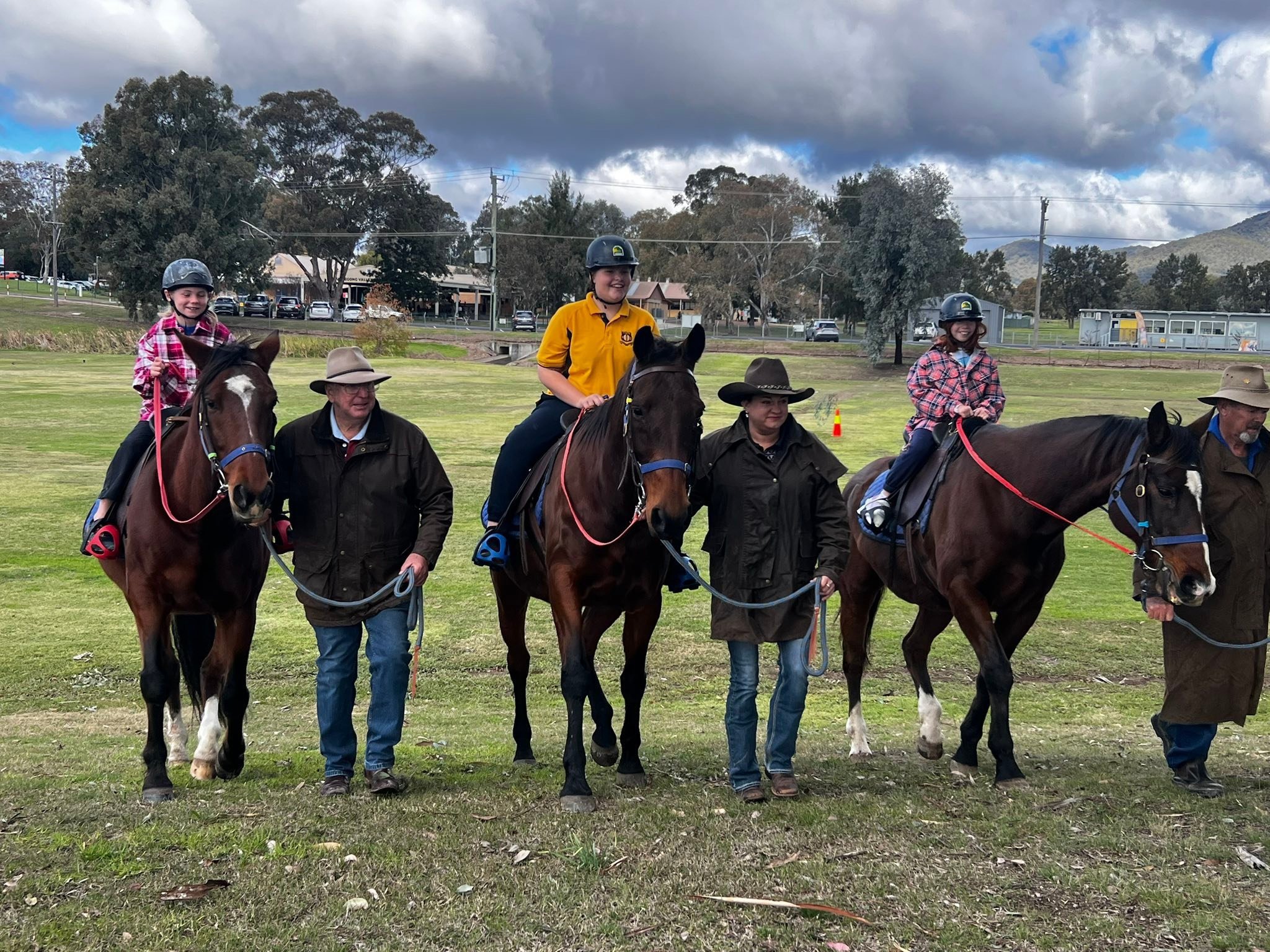 Children riding horses in an outdoor field with adults walking alongside, under a cloudy sky.